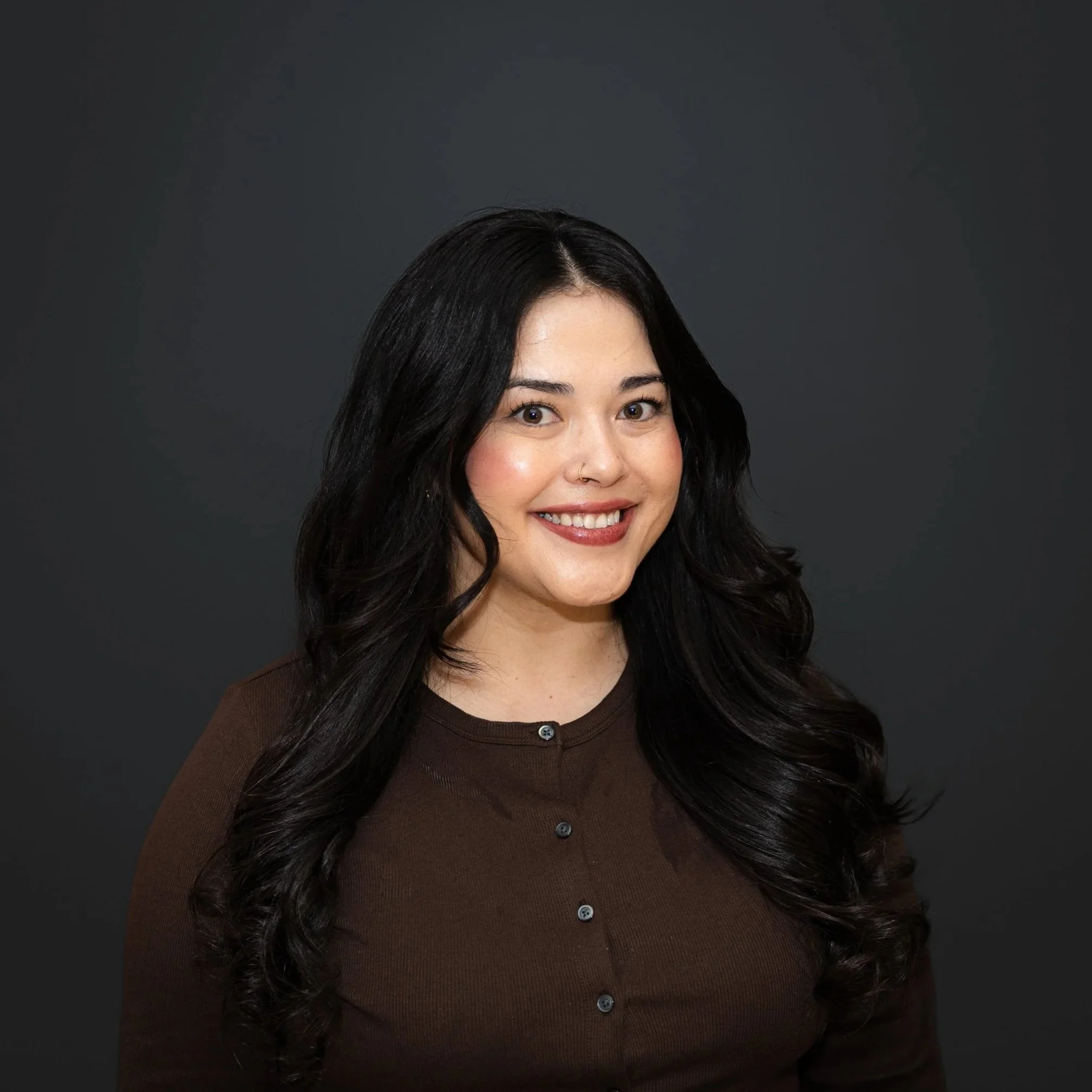 Portrait of a woman with dark hair and highlights, wearing a black collared shirt, against a dark background.