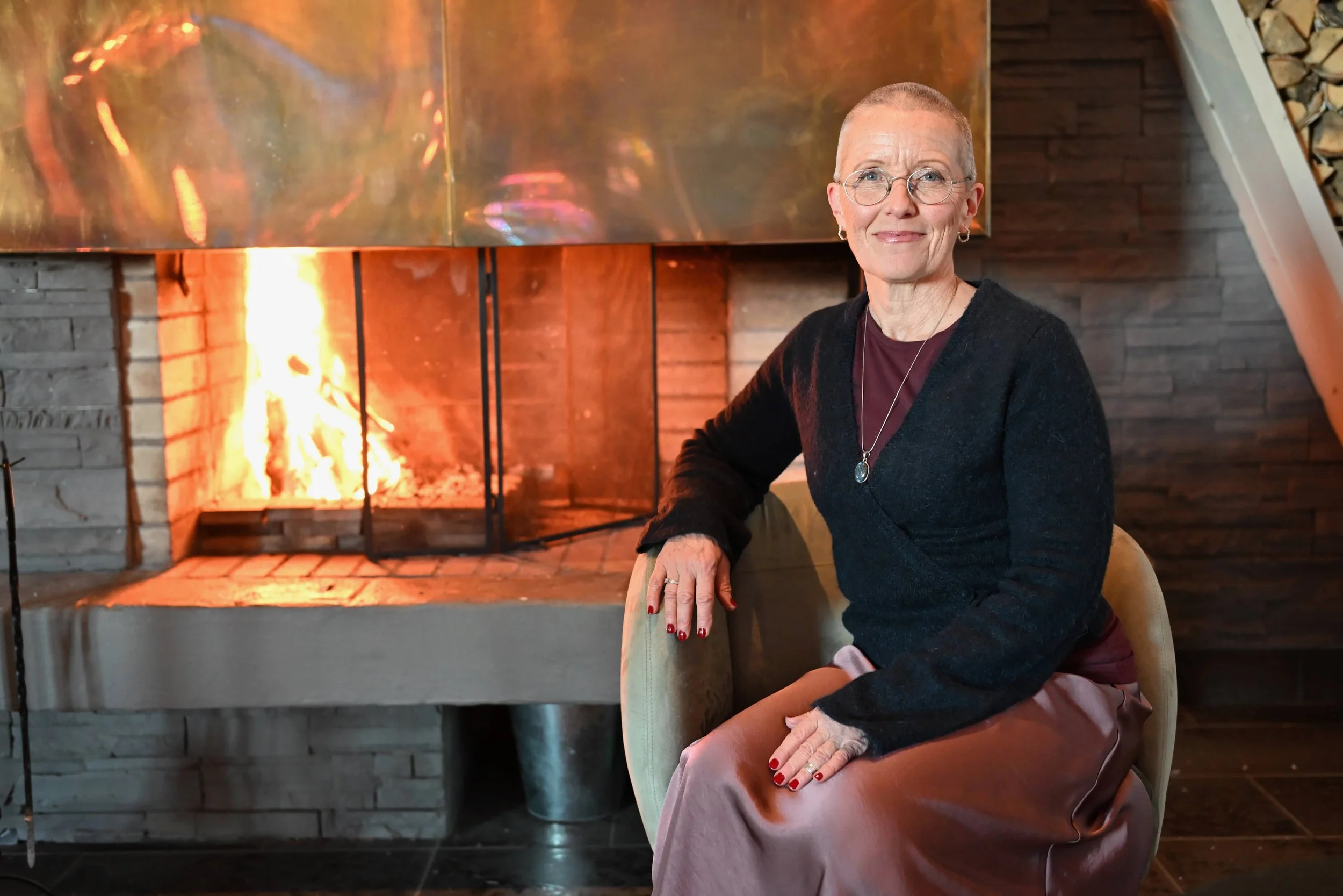 An elderly woman sitting in front of a lit fireplace with a fire burning inside, wearing glasses and a black sweater.