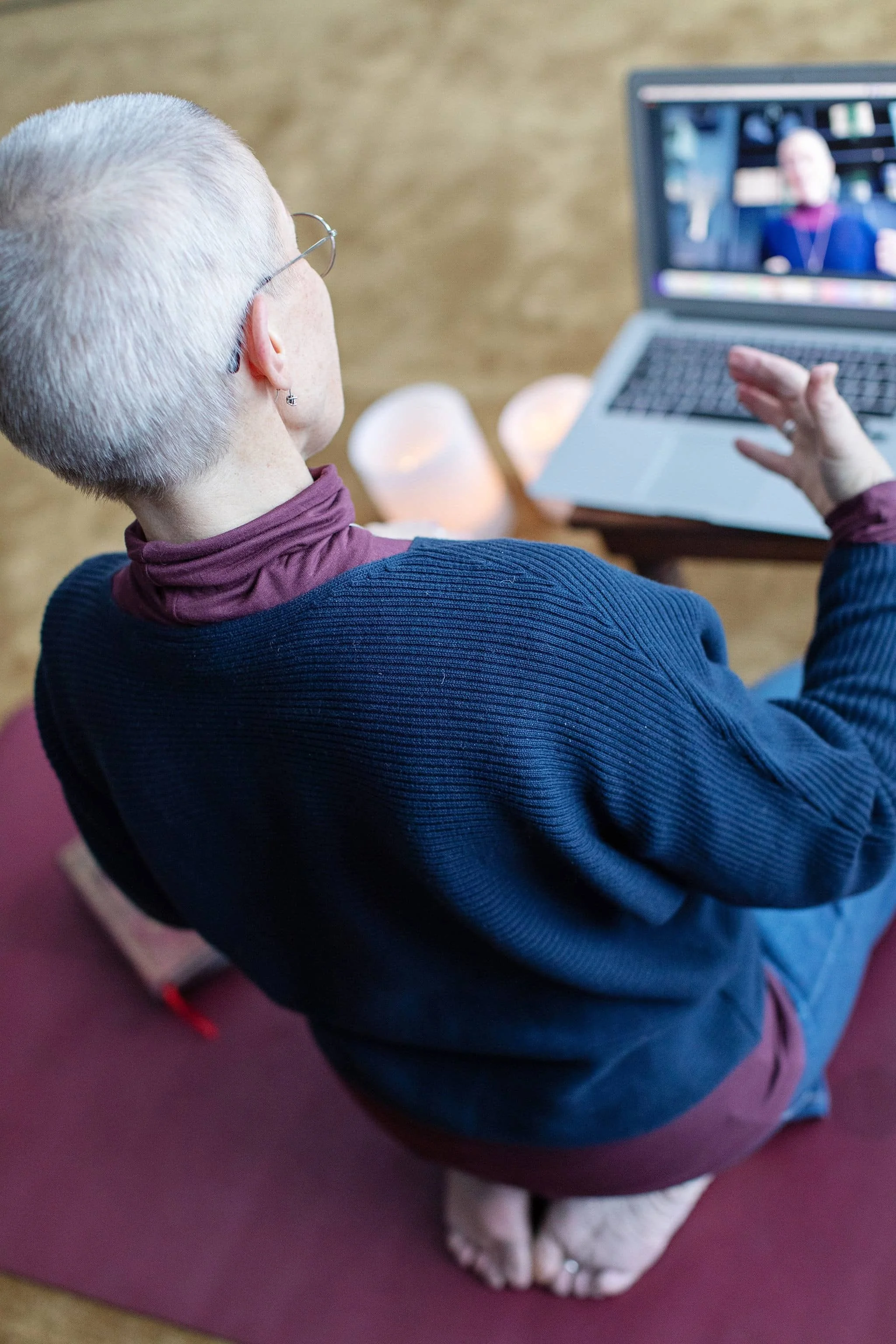A person with short gray hair is sitting on a gym mat, participating in an online yoga class while on their knees, with half their body visible. They are wearing glasses and a dark blue sweater, and are facing a laptop with a video of an instructor.