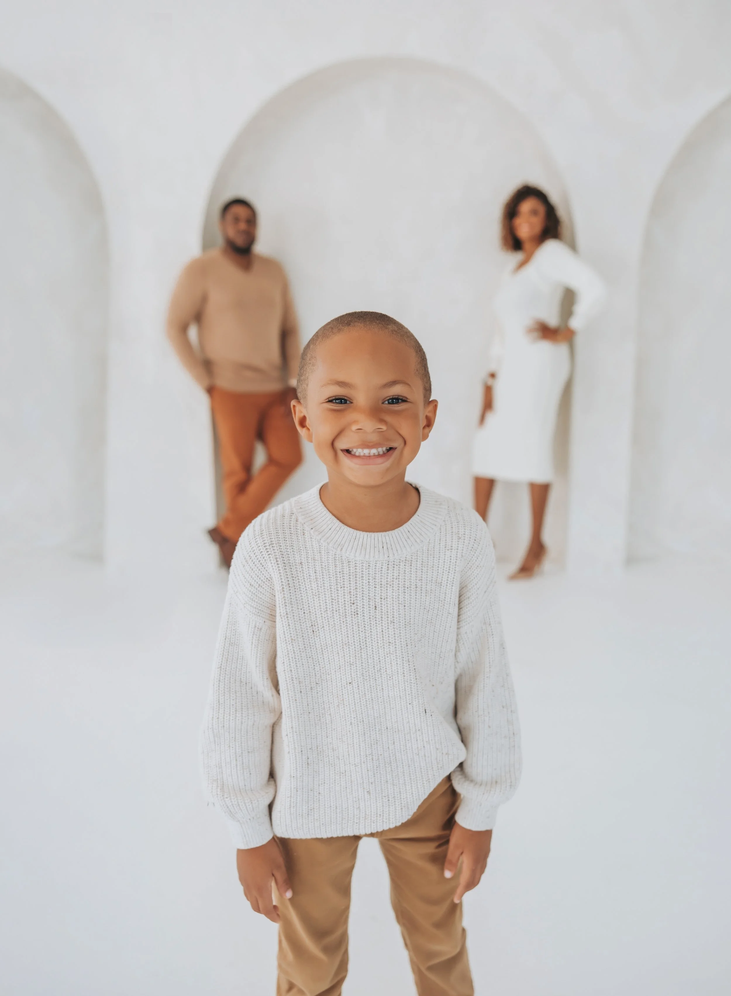 A young boy smiling in the foreground, with a man and woman standing in the background, all in a white, minimalist setting.