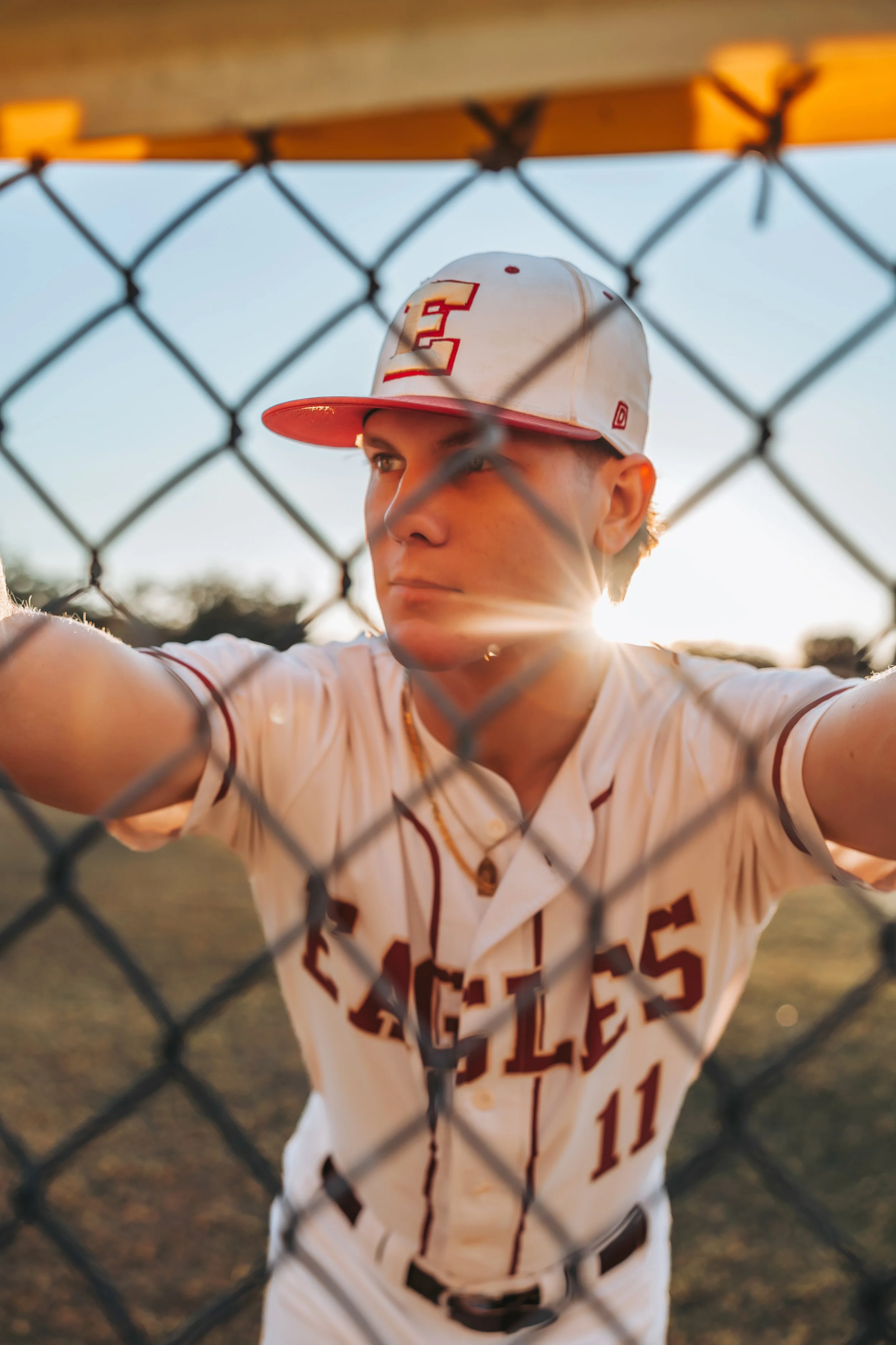 A young male baseball player in a white uniform with red accents, standing behind a chain-link fence during sunset, wearing a baseball cap with a red and yellow emblem.