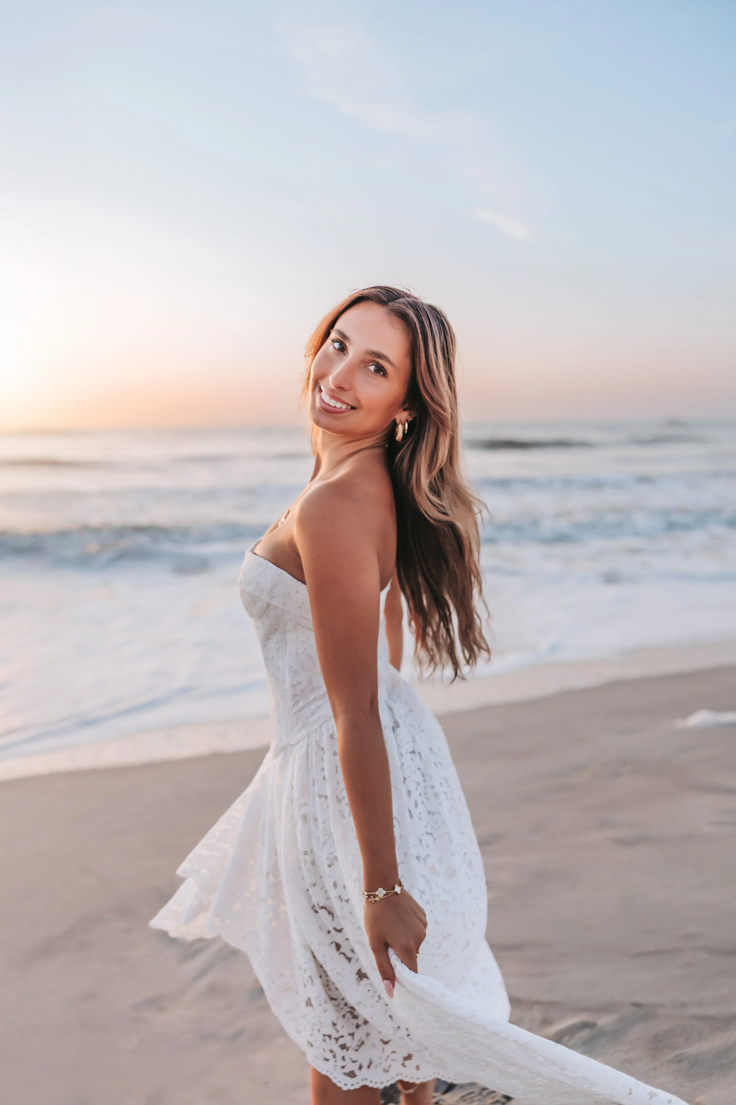 A woman in a white lace dress smiling on the beach during sunrise or sunset with the ocean in the background.