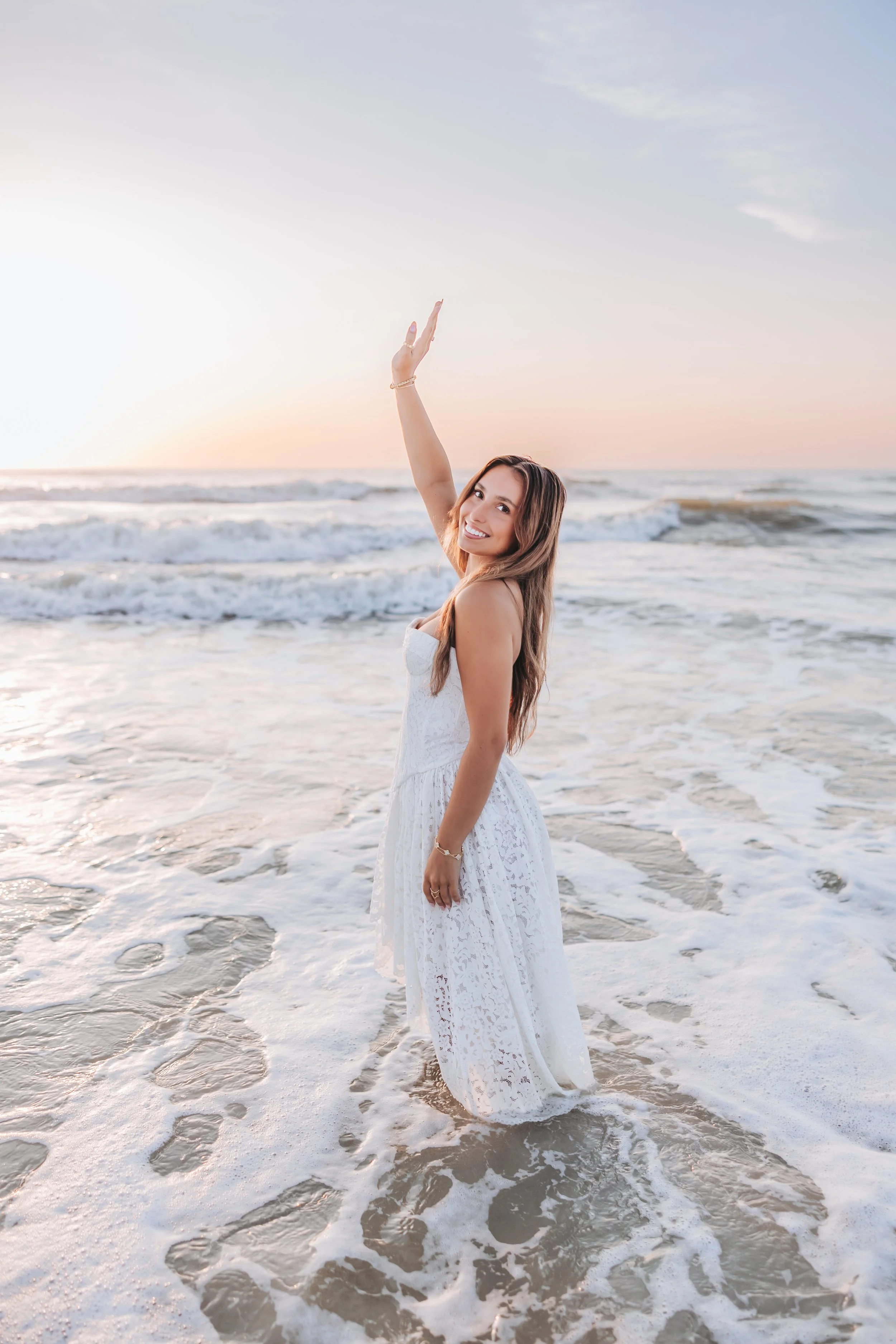 A woman in a white dress standing in the ocean with waves around her and smiling at the camera during sunset.