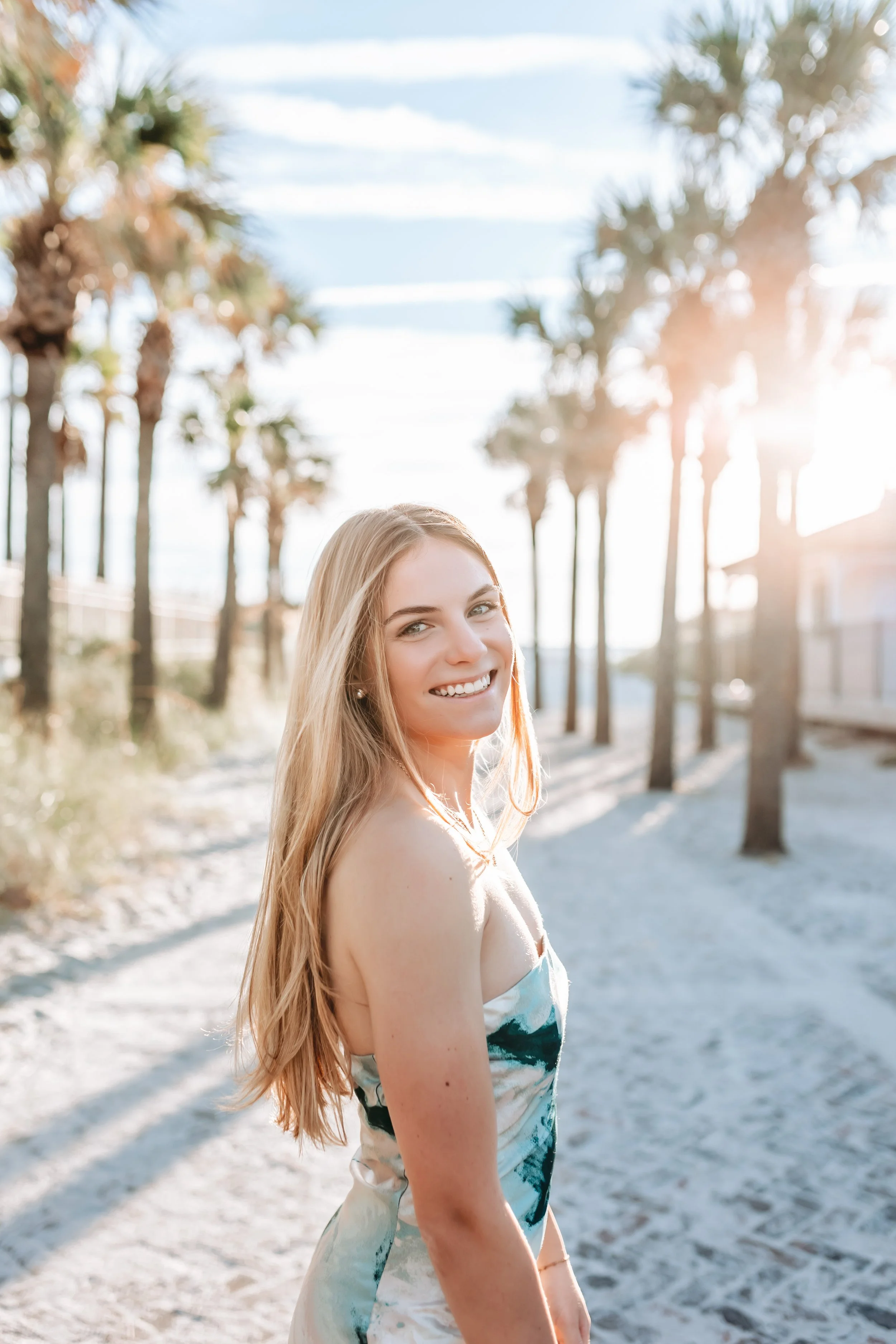 Young woman with long blonde hair smiling at the camera on a sunny beach with palm trees in the background.