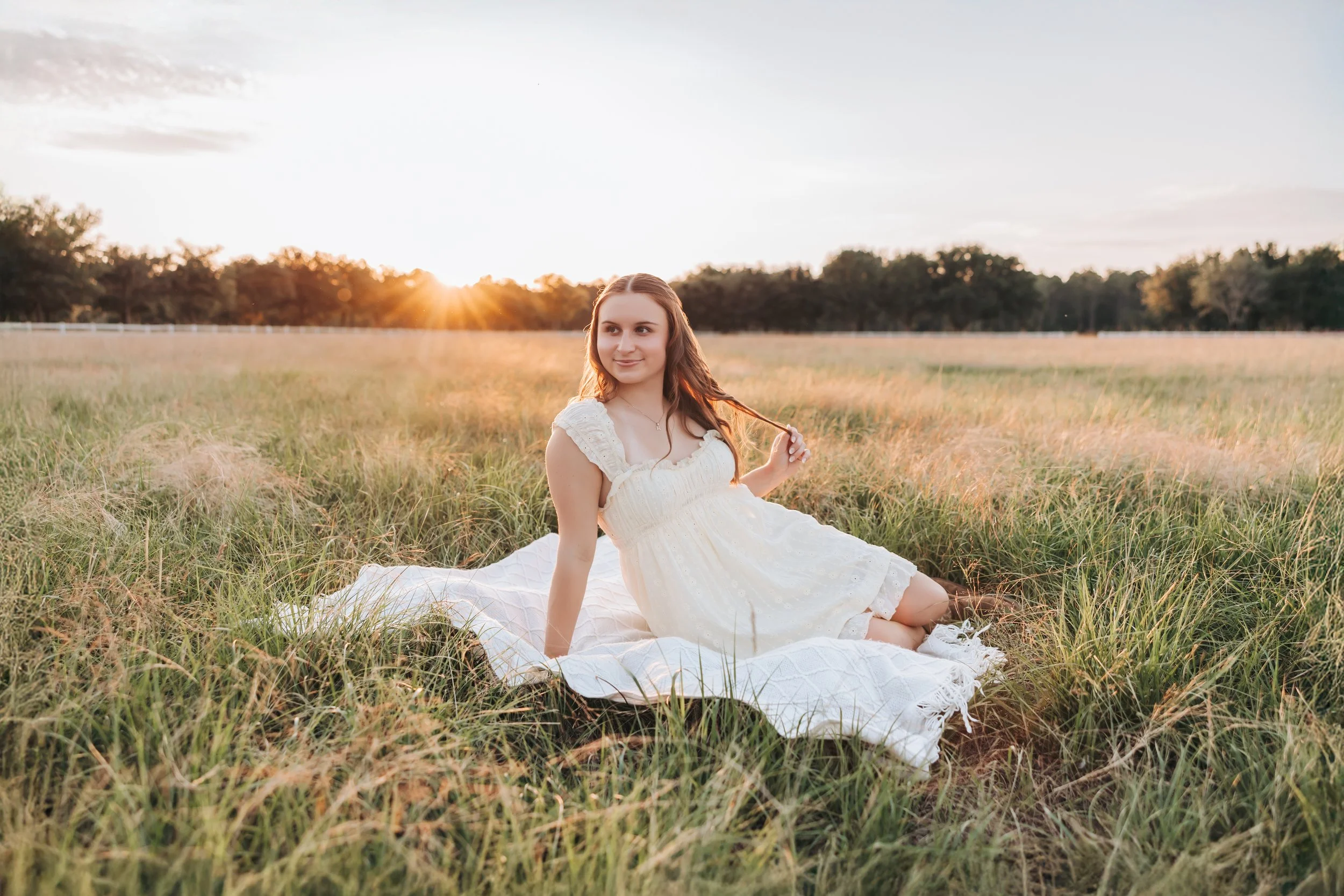 A young woman in a white dress sitting on a blanket in a grassy field during sunset.