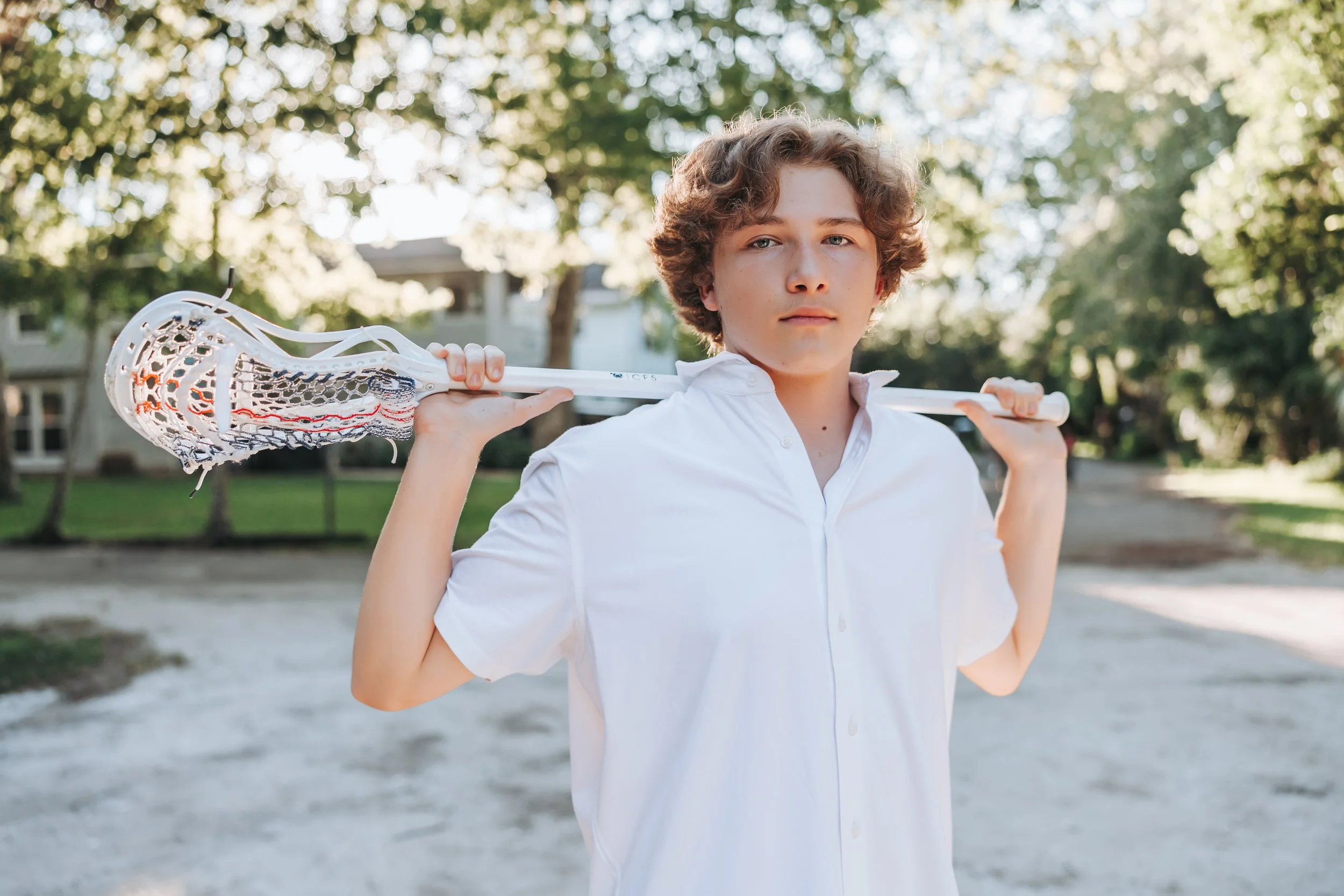 Young person holding lacrosse stick over shoulders outdoors, wearing a white shirt, with trees and sunlight in the background.