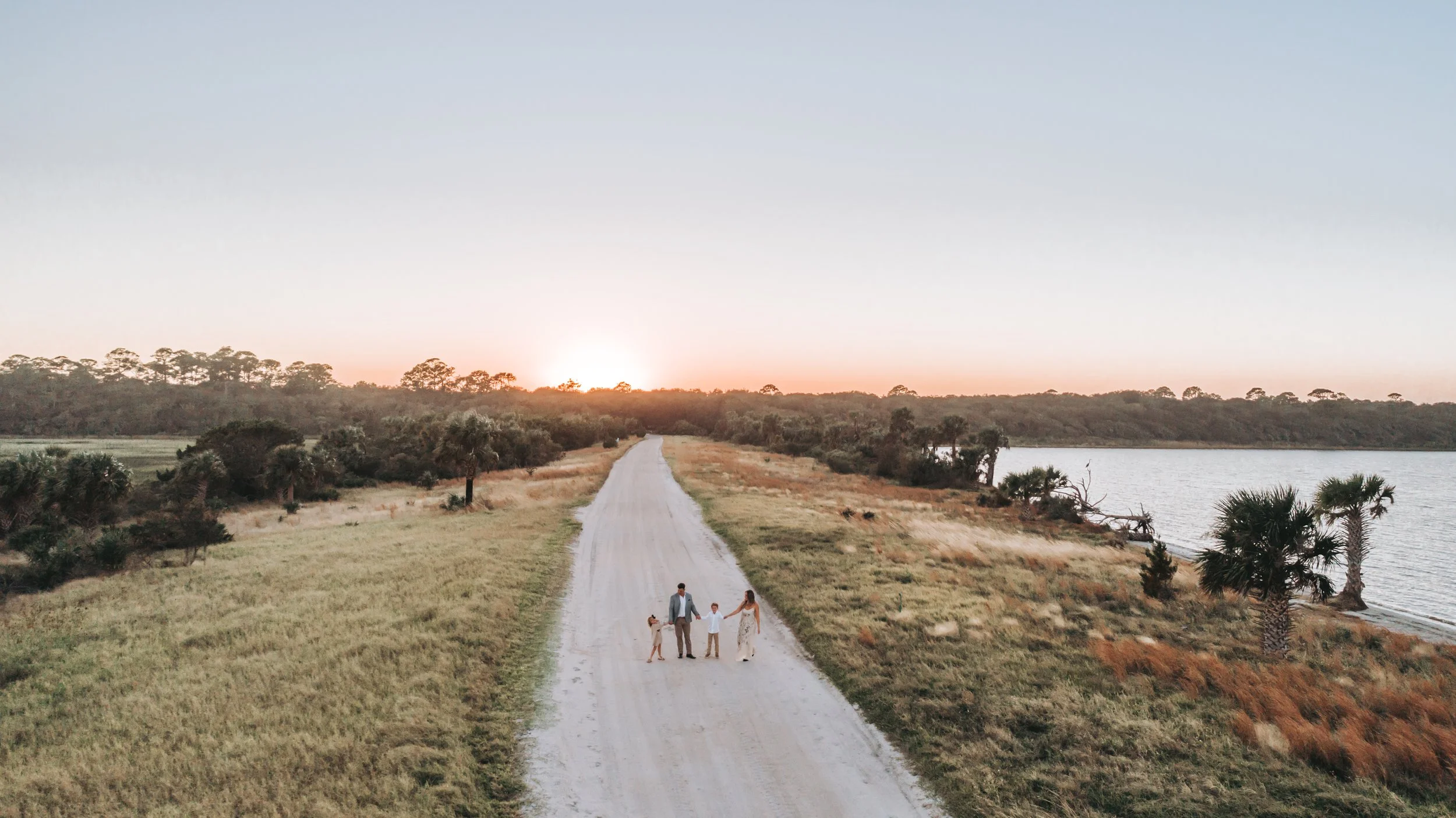 A family of four walking hand-in-hand on a dirt path along a lakeside during sunset, surrounded by trees and open grassland.