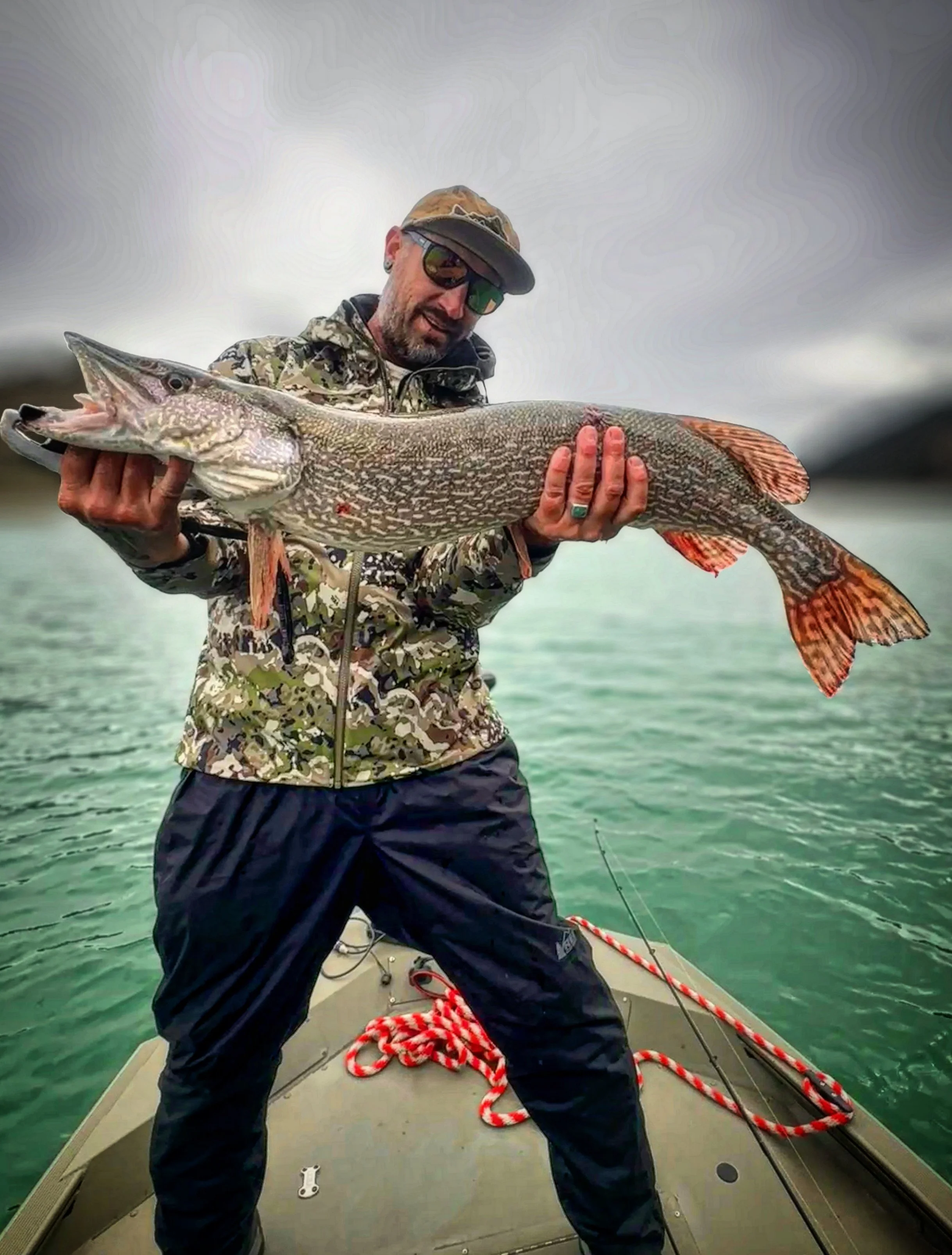 Man in camouflage jacket and dark pants holding large fish on boat, over cloudy water.