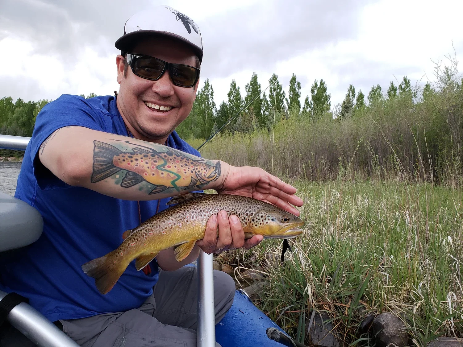 Man sitting in a boat holding a fish with a remote spot of water and trees in the background. The man is wearing a blue shirt, sunglasses, and a cap.