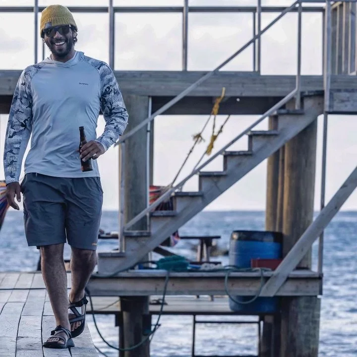 Man with a beard, wearing a yellow beanie, sunglasses, a long-sleeved outdoor shirt, shorts, and sandals, standing on a dock by the ocean, smiling and holding a bottle.