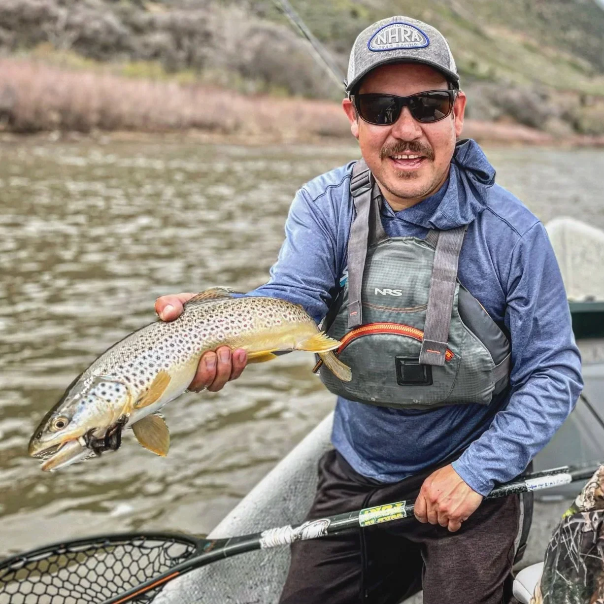Man holding a large fish on a boat, wearing sunglasses, a gray hat, and outdoor gear, with a river and hilly landscape in the background.