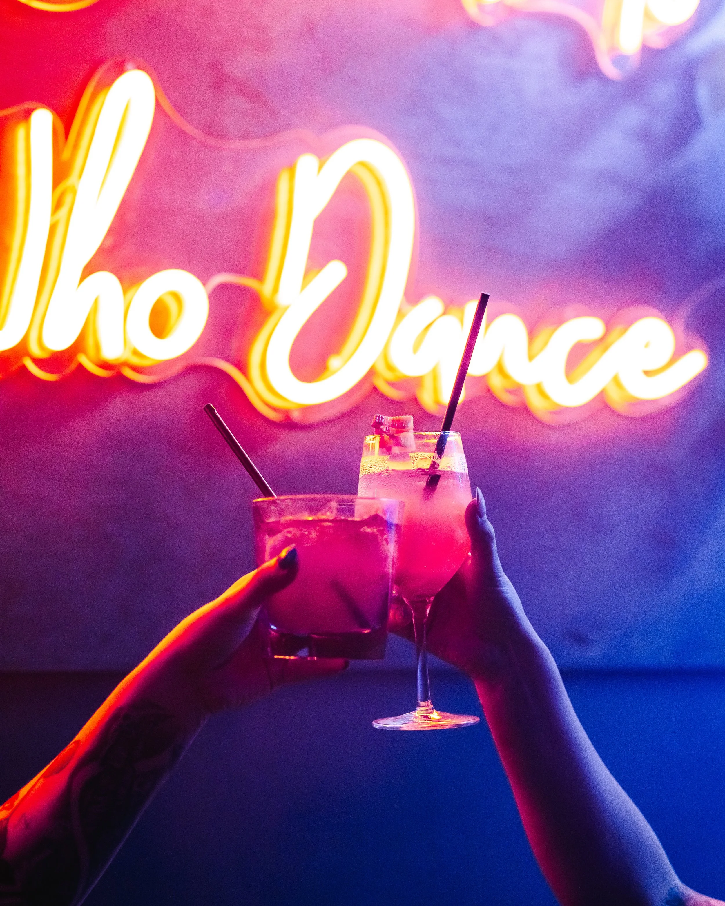 Two hands, one with tattoos, holding pink cocktails with straws in front of a neon sign that reads 'Illo Dance' in colorful lights.