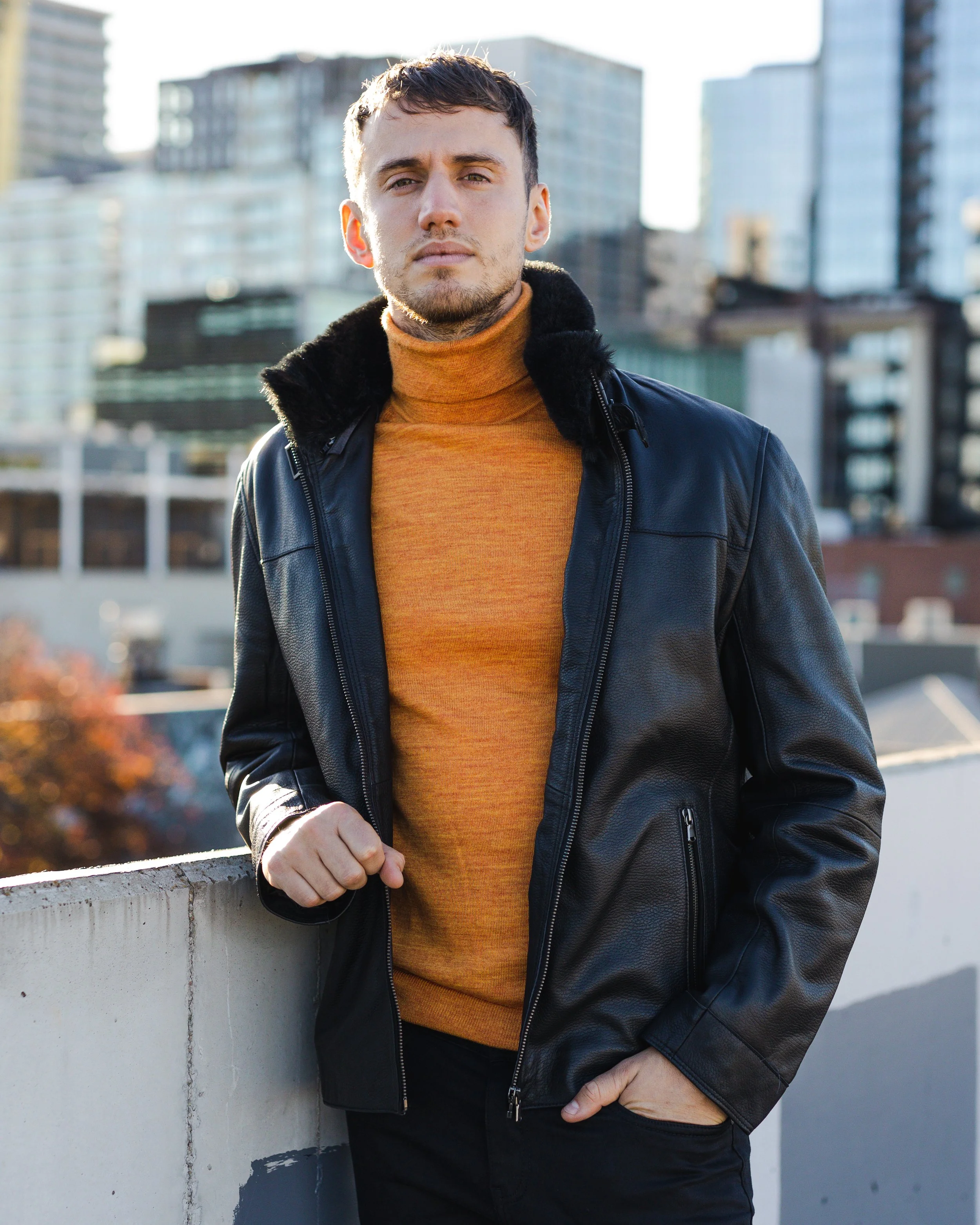 A young man in a black leather jacket and orange turtleneck standing outdoors on a city rooftop with tall buildings in the background.
