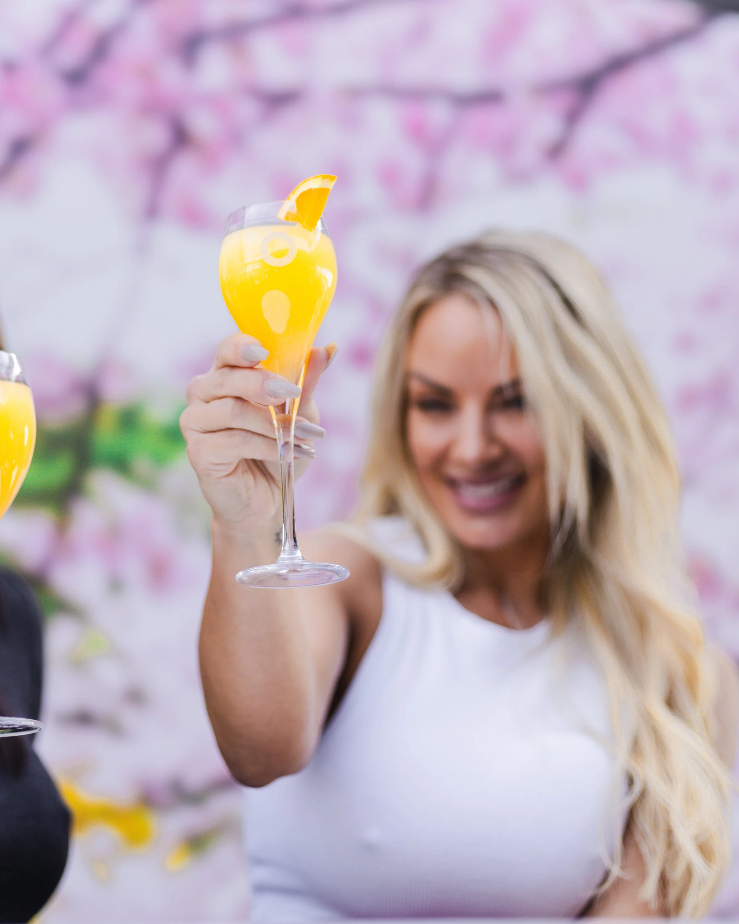 A woman with long blonde hair holding a glass of mimosa with an orange slice garnish, smiling at a gathering