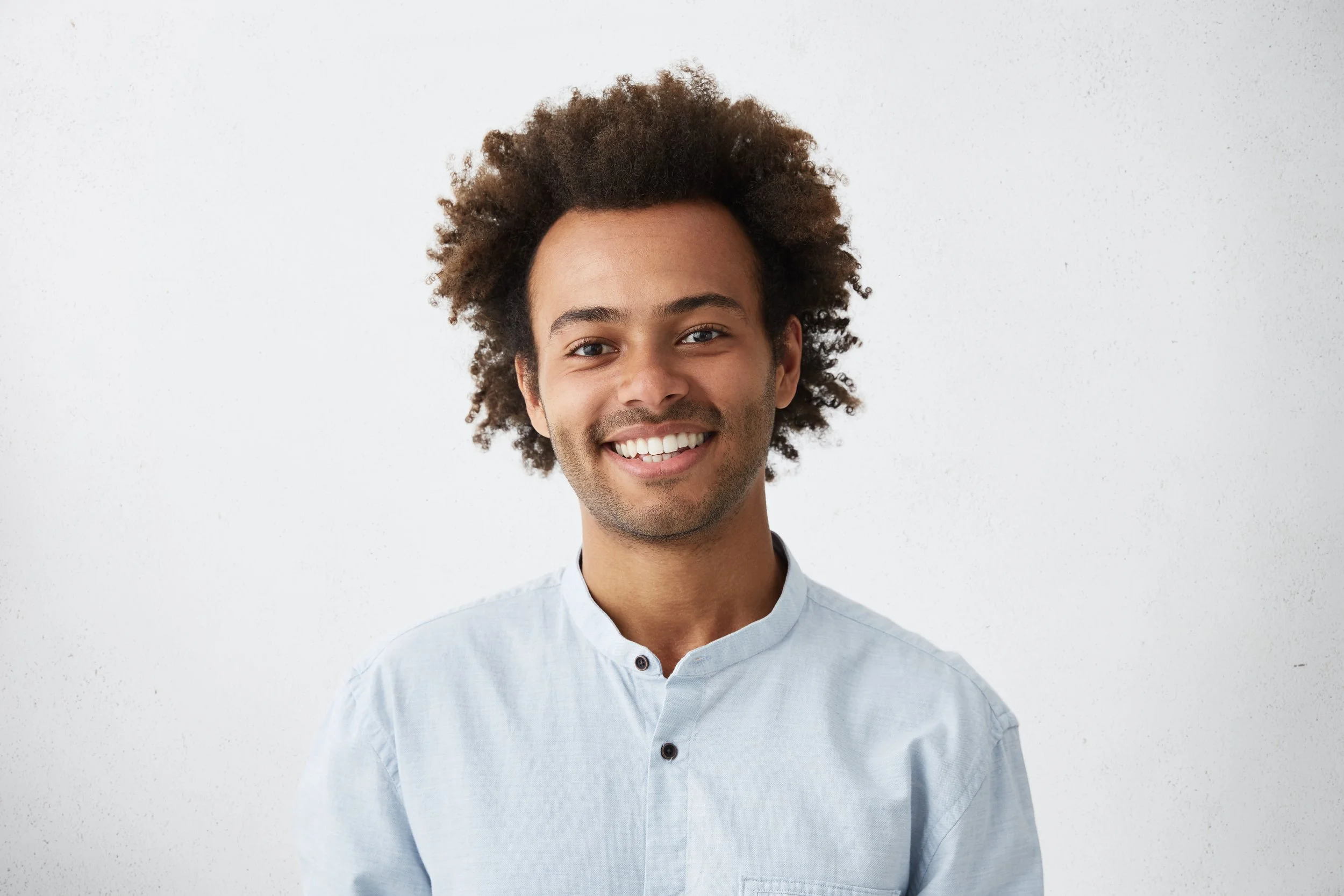 Smiling young man with curly hair wearing a light blue shirt against a plain white wall.