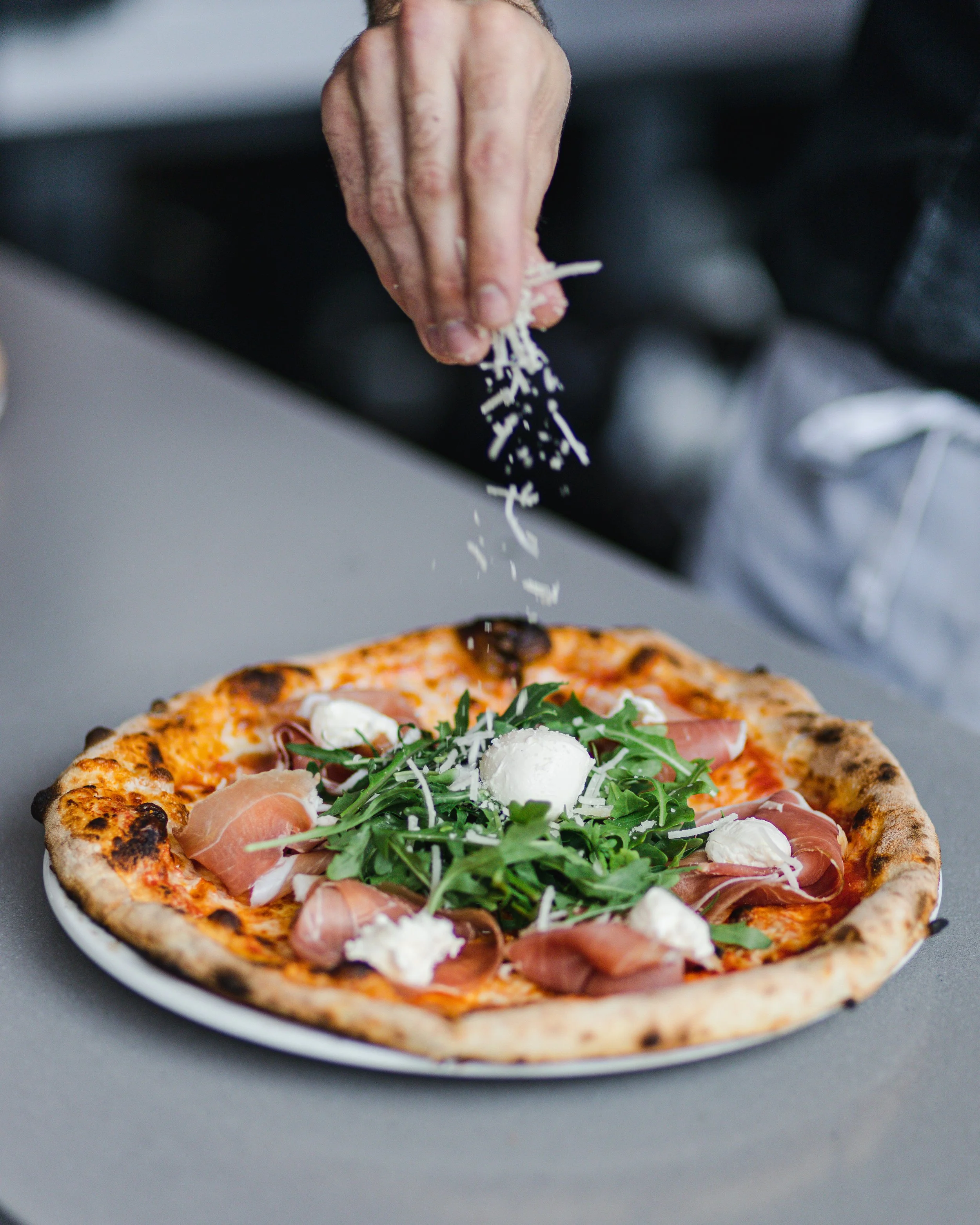 Person grating cheese onto a pizza topped with prosciutto, arugula, mozzarella, and tomato sauce.