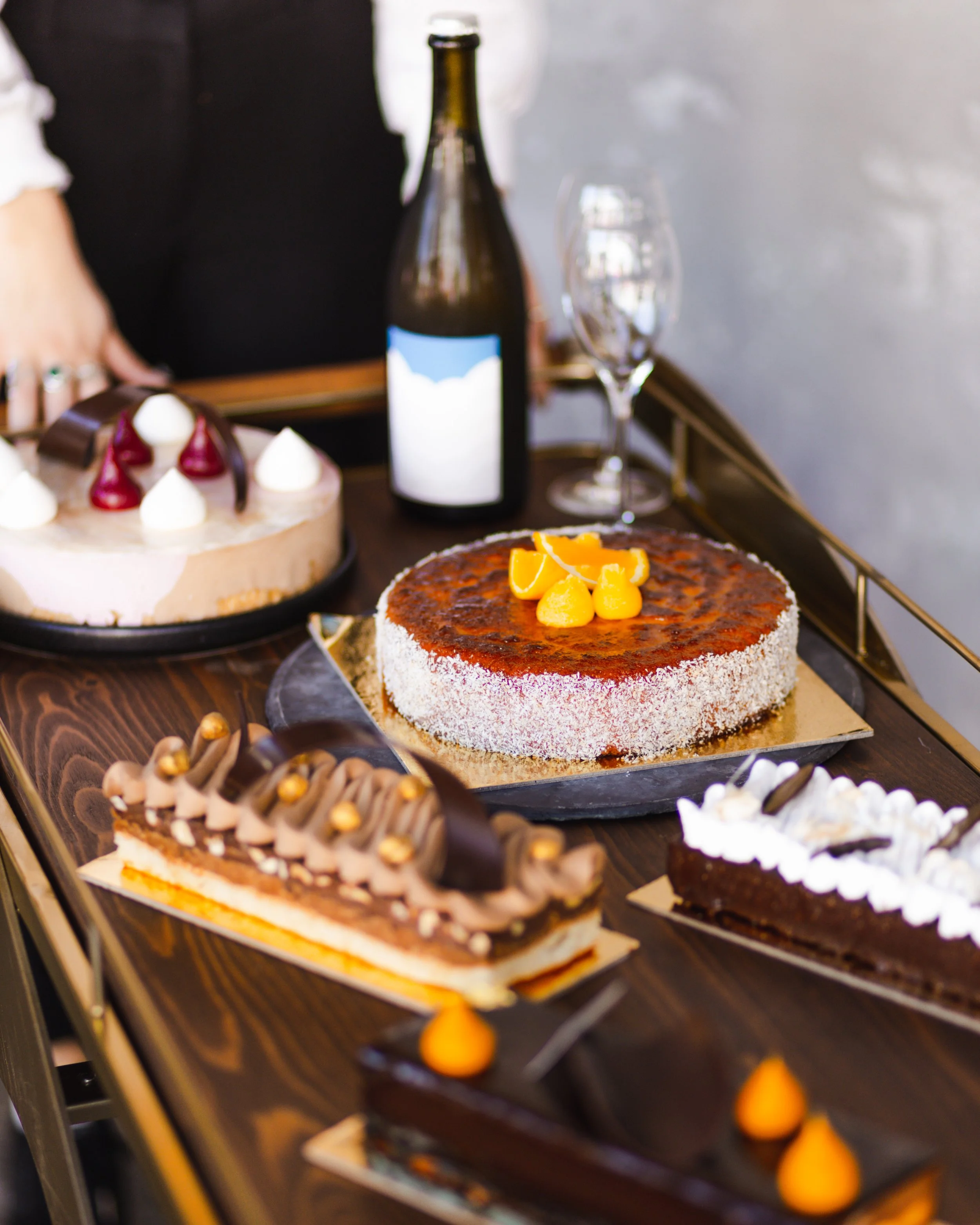 Assorted cakes on a table, including a round cake with orange slices on top, a chocolate dessert with gold accents, a vanilla cake with cherries and chocolate, and a rectangular chocolate cake with dollops of cream and orange decorations, with a bott