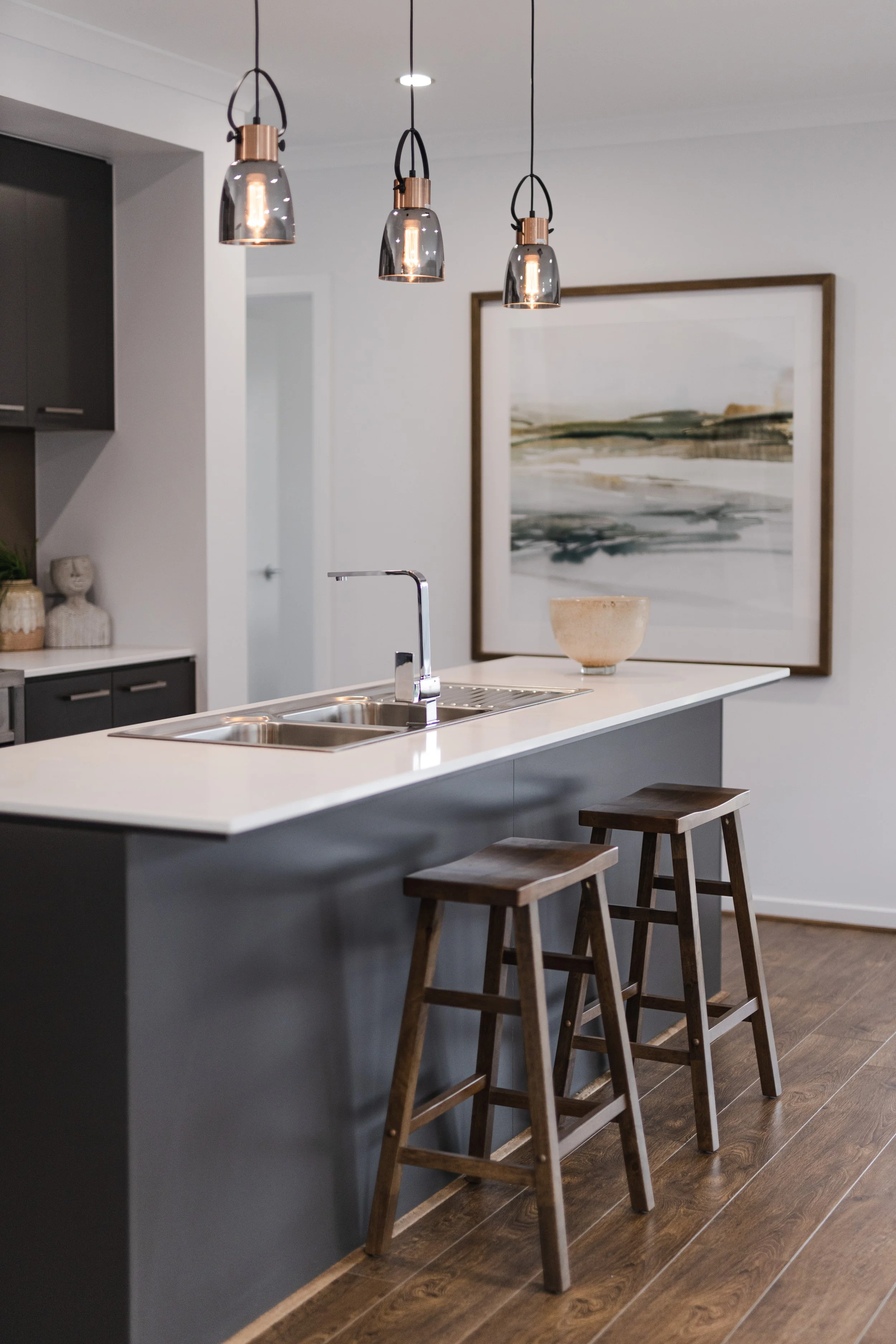 Modern kitchen with pendant lights, a white countertop island, wooden stools, and a large framed abstract painting on the wall.