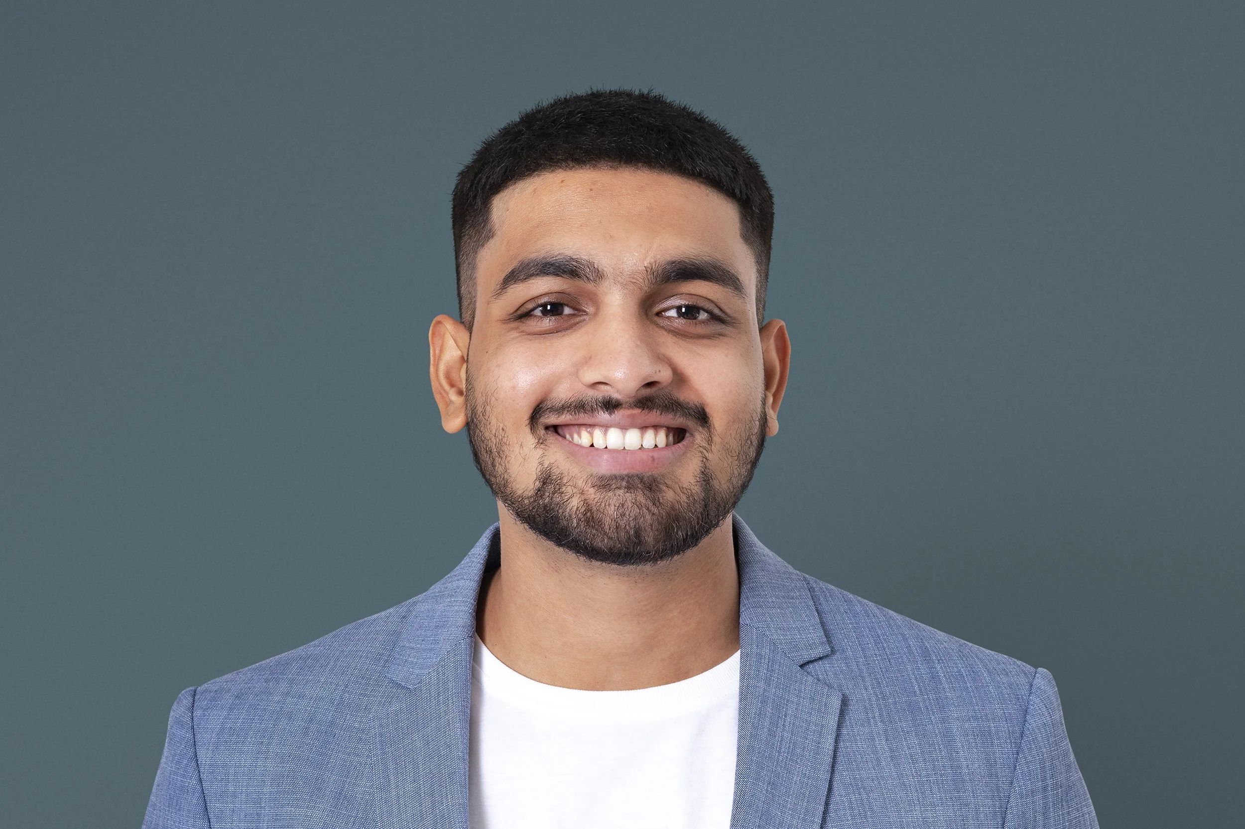 A smiling man with short dark hair and a beard, wearing a light blue blazer over a white t-shirt, standing against a solid green background.