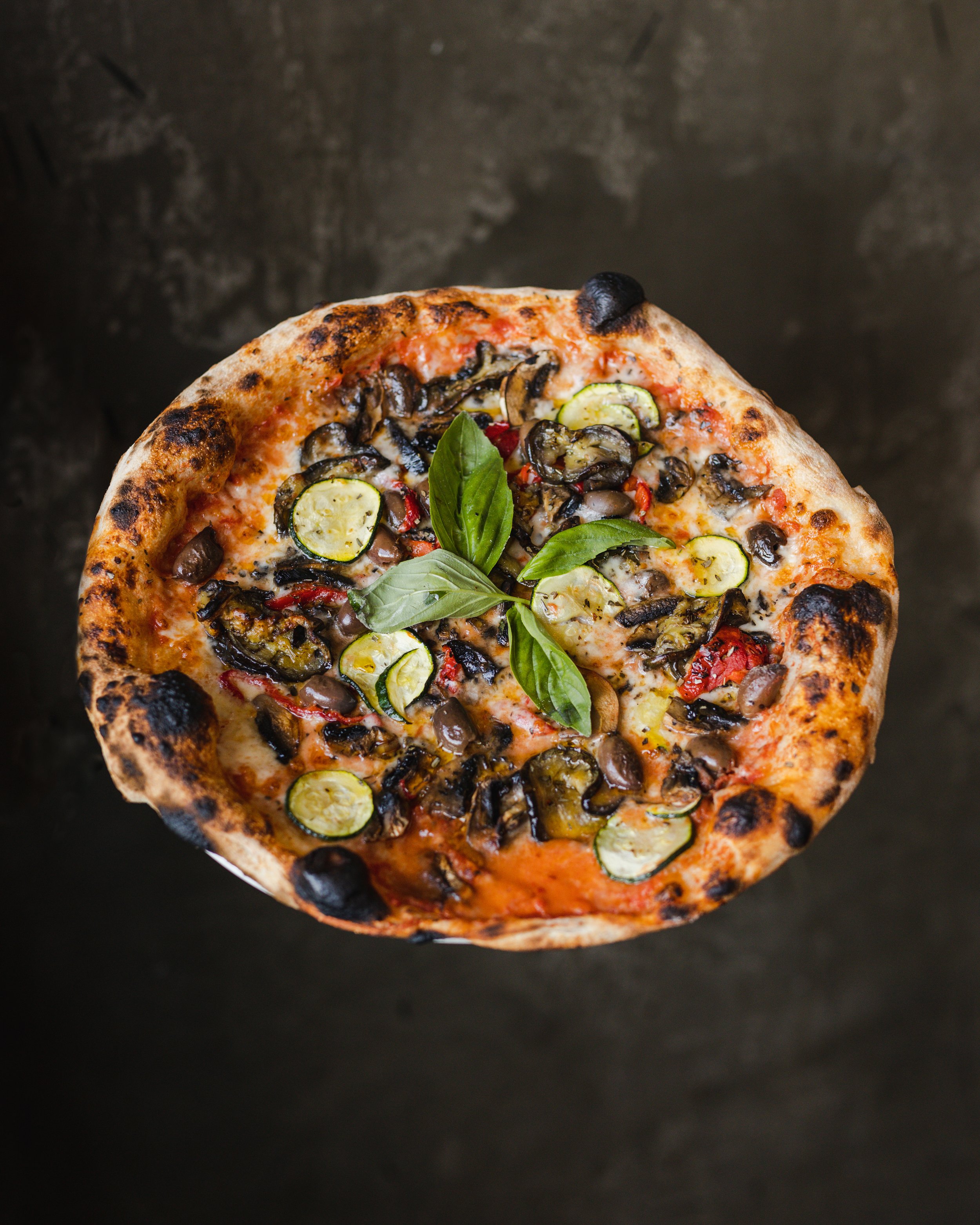 A top view of a pizza topped with sliced zucchini, mushrooms, red peppers, and fresh basil leaves on a dark surface.