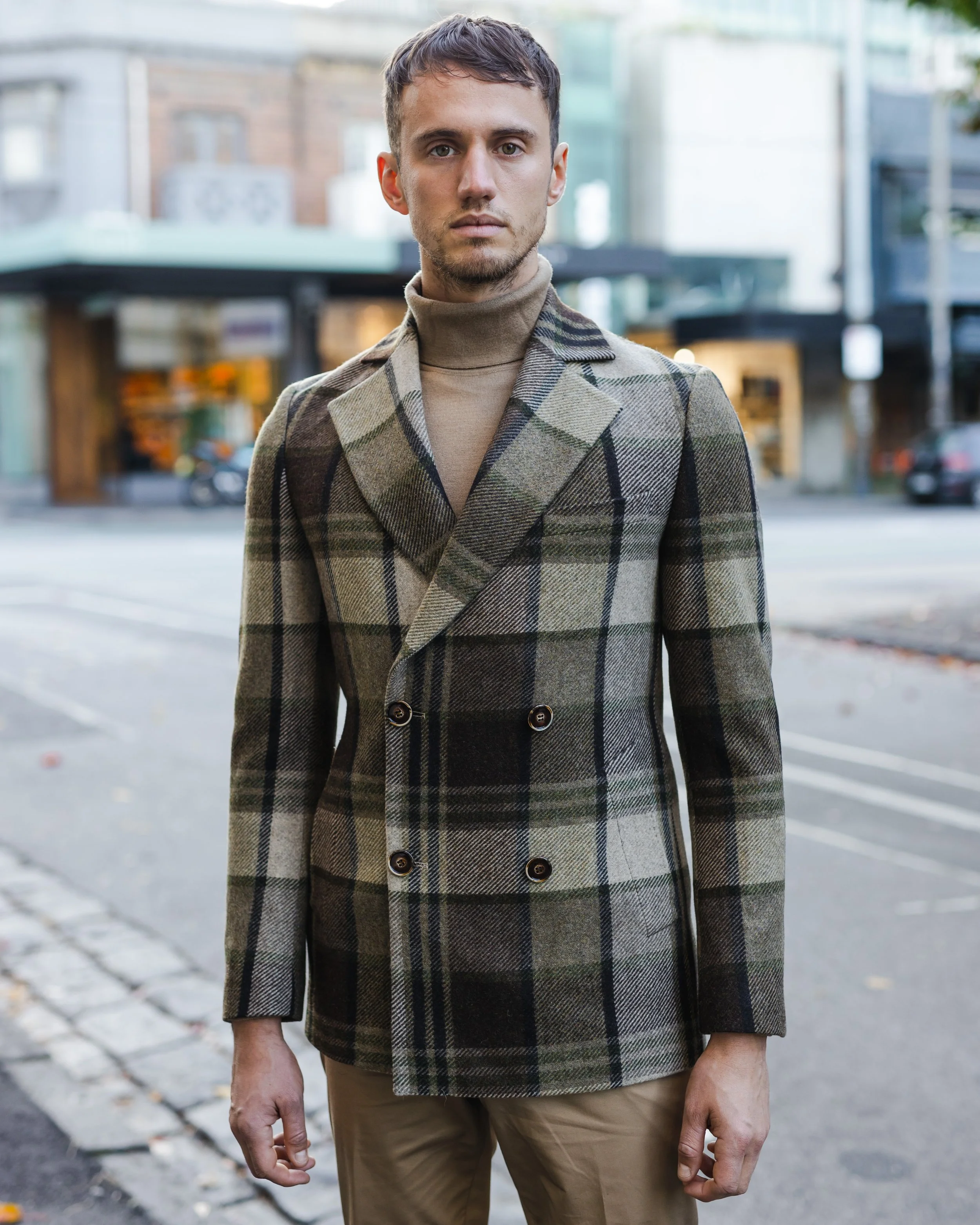 A man standing outdoors on a city street, wearing a plaid blazer and a turtleneck sweater.