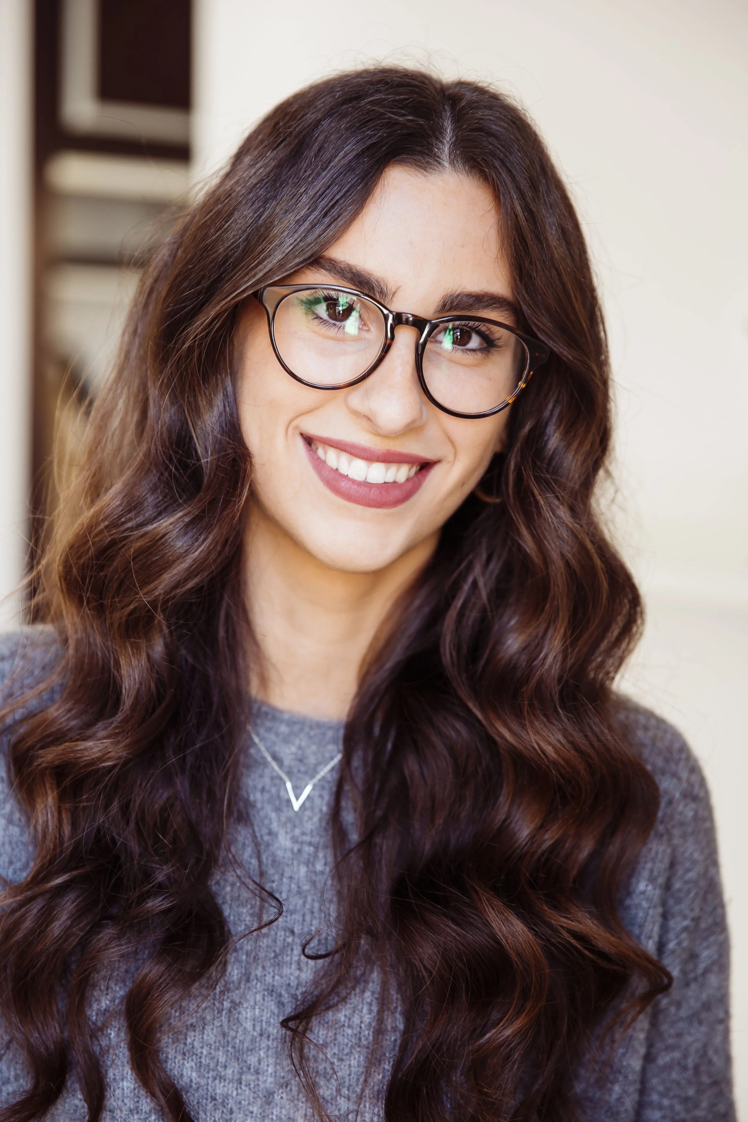 A young woman with long, wavy brown hair and glasses, smiling at the camera, wearing a gray top and a delicate necklace.