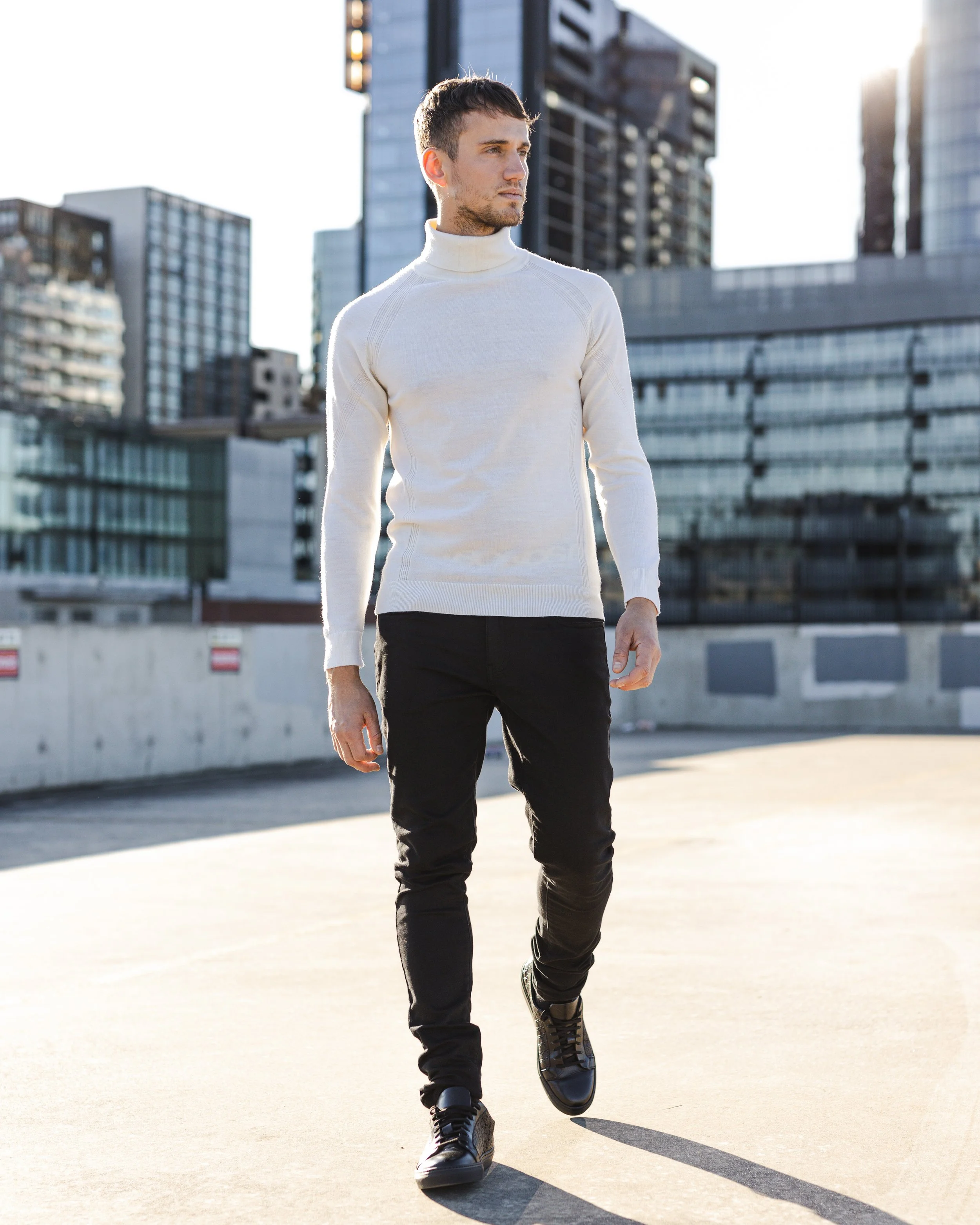 A man walking outdoors in an urban area with modern glass buildings in the background, wearing a white turtleneck sweater, black pants, and black shoes.