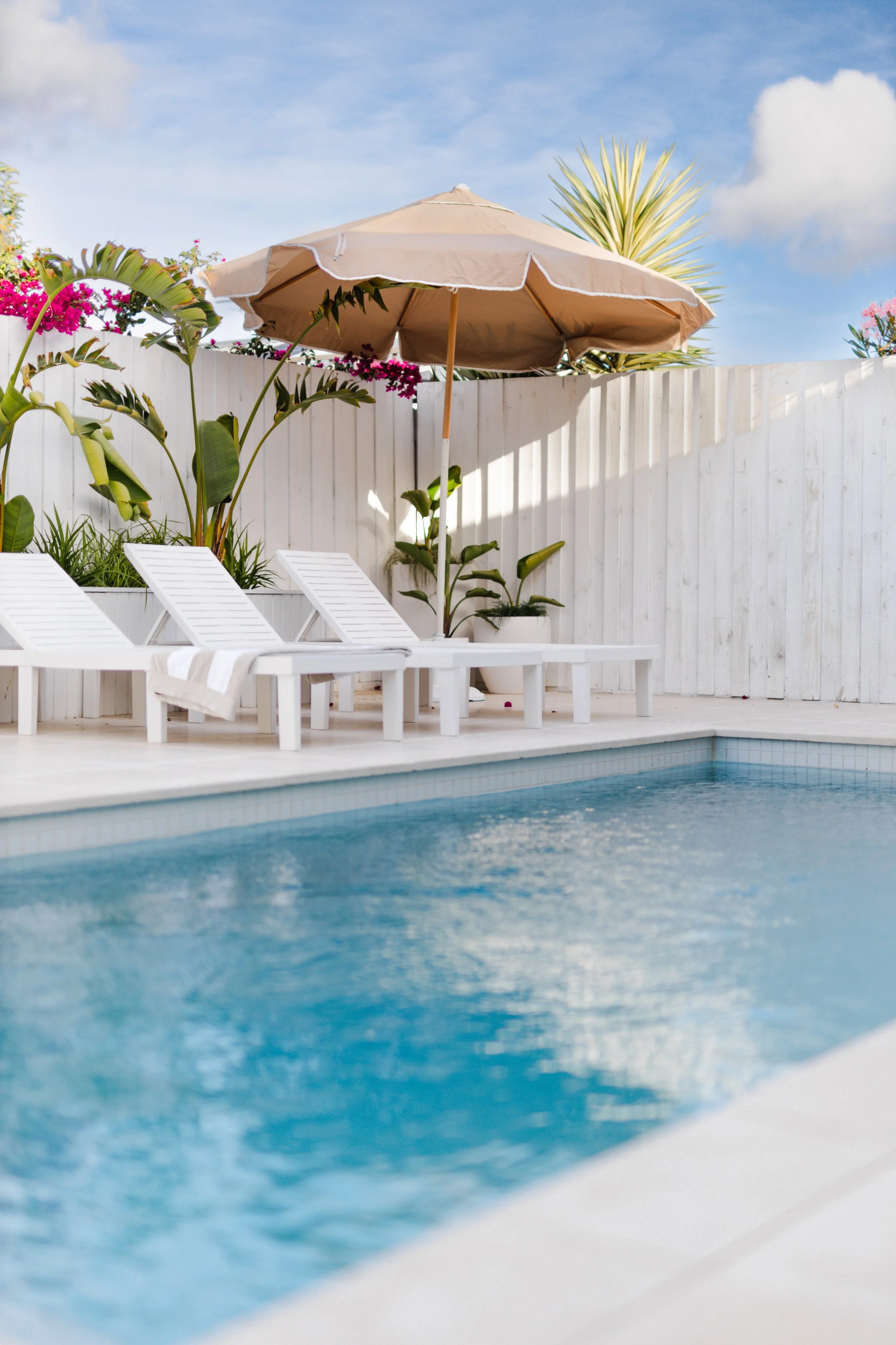 A backyard swimming pool with white poolside lounge chairs, a beige umbrella, and tropical plants along a white wooden fence under a blue sky with clouds.