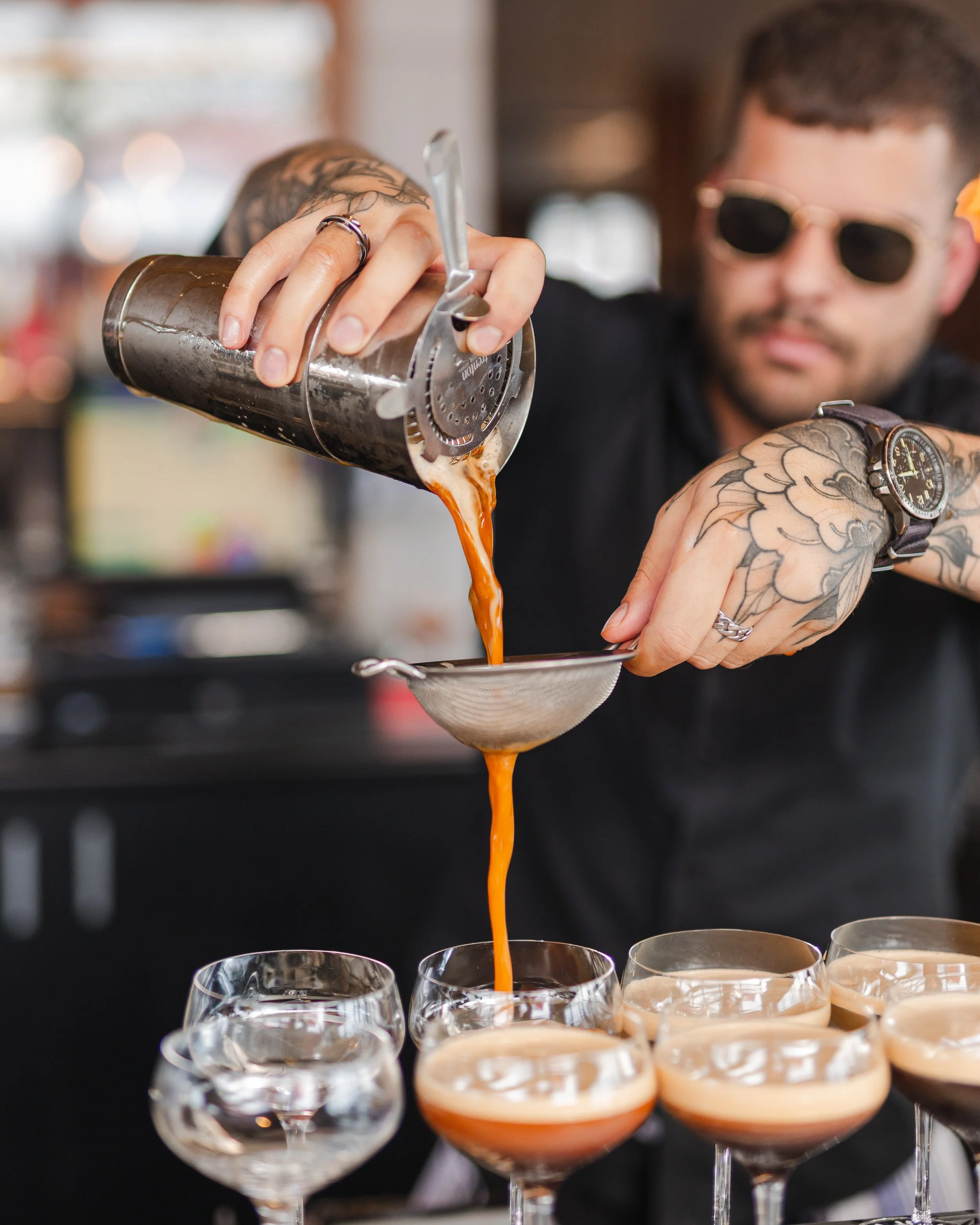 A bartender with tattoos and sunglasses is pouring a cocktail from a shaker through a strainer into a glass. Multiple cocktail glasses are lined up in front.