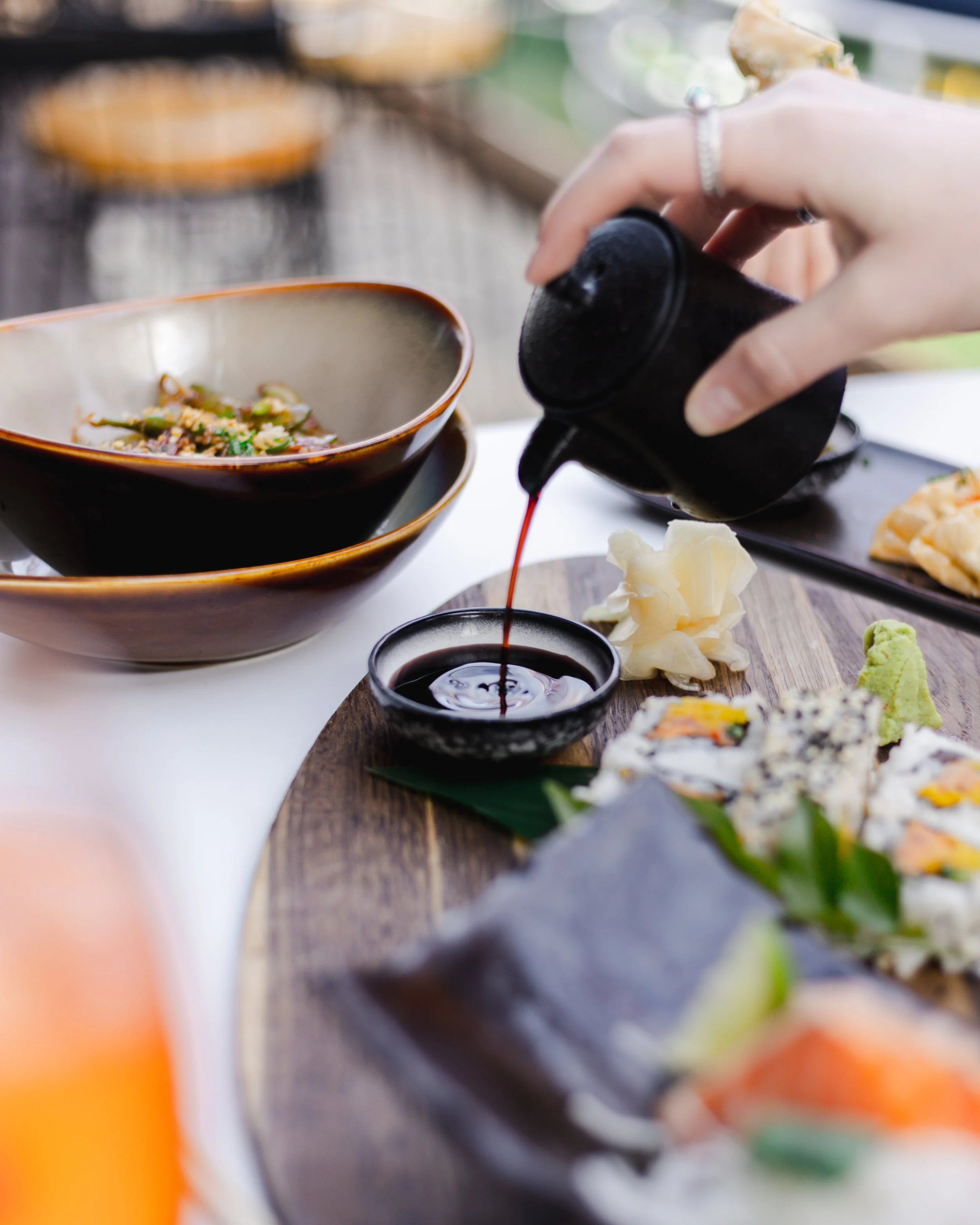 Person pouring soy sauce onto sushi and sashimi on a wooden platter at a restaurant.