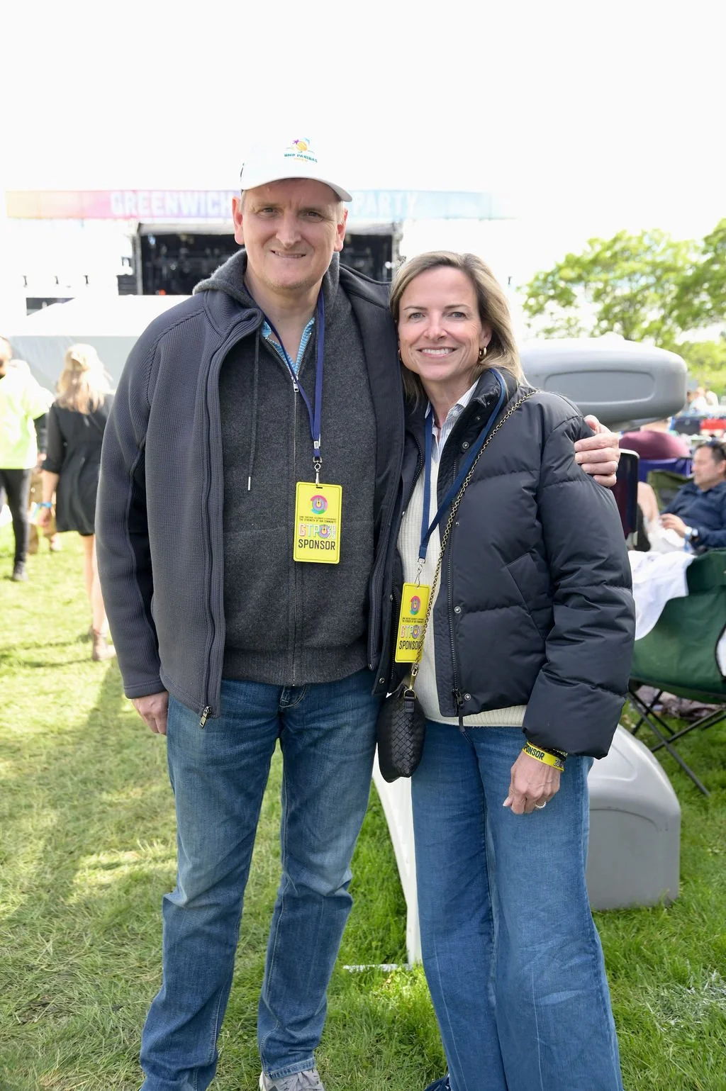 A man and a woman stand together outdoors at a festival or outdoor event, smiling at the camera. They are wearing jackets and have event sponsor badges hanging around their necks. In the background, there is a stage with a banner that reads 'Greenwic