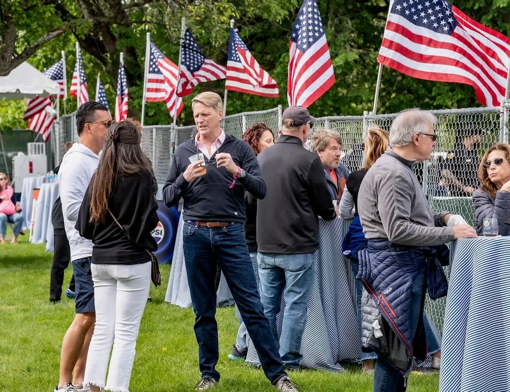 People gathered outdoors at a patriotic event with American flags, some standing near a fence, others sitting at tables, during daytime.