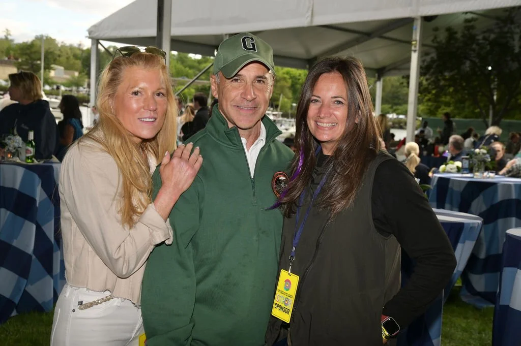 Three people smiling at an outdoor event under a tent, with tables and other guests in the background.