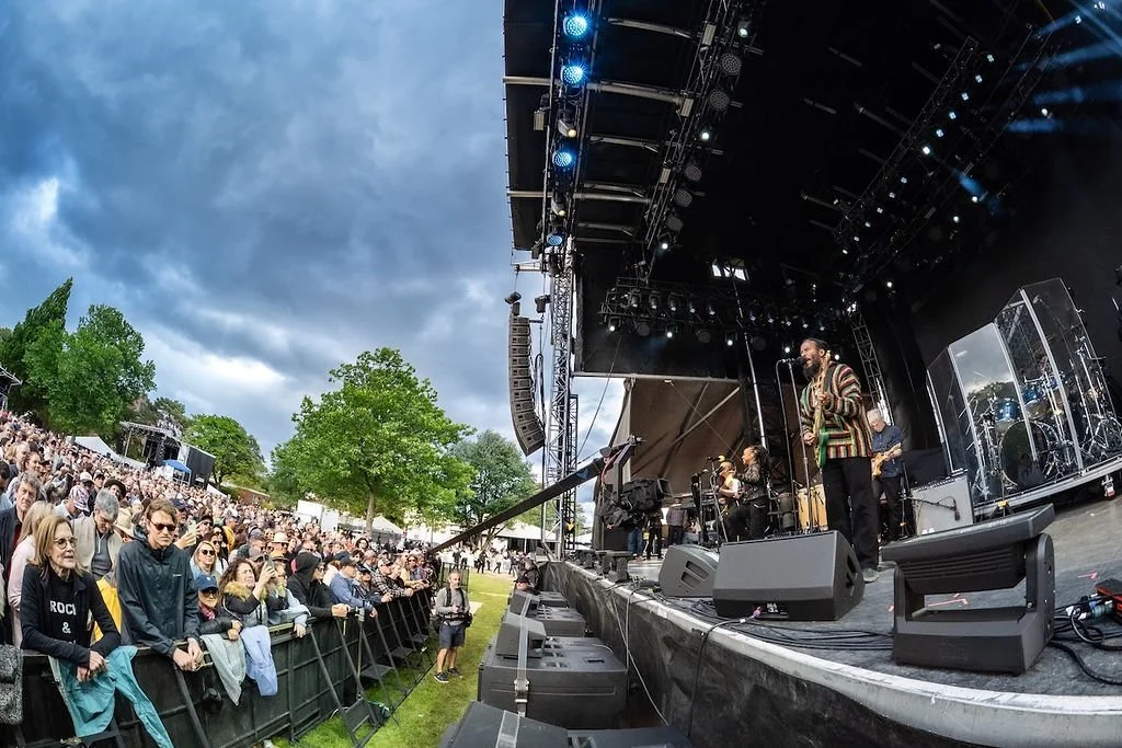 A live outdoor concert shows a band performing on stage with the audience standing and watching. The stage has lights and musical equipment, and the sky is cloudy.