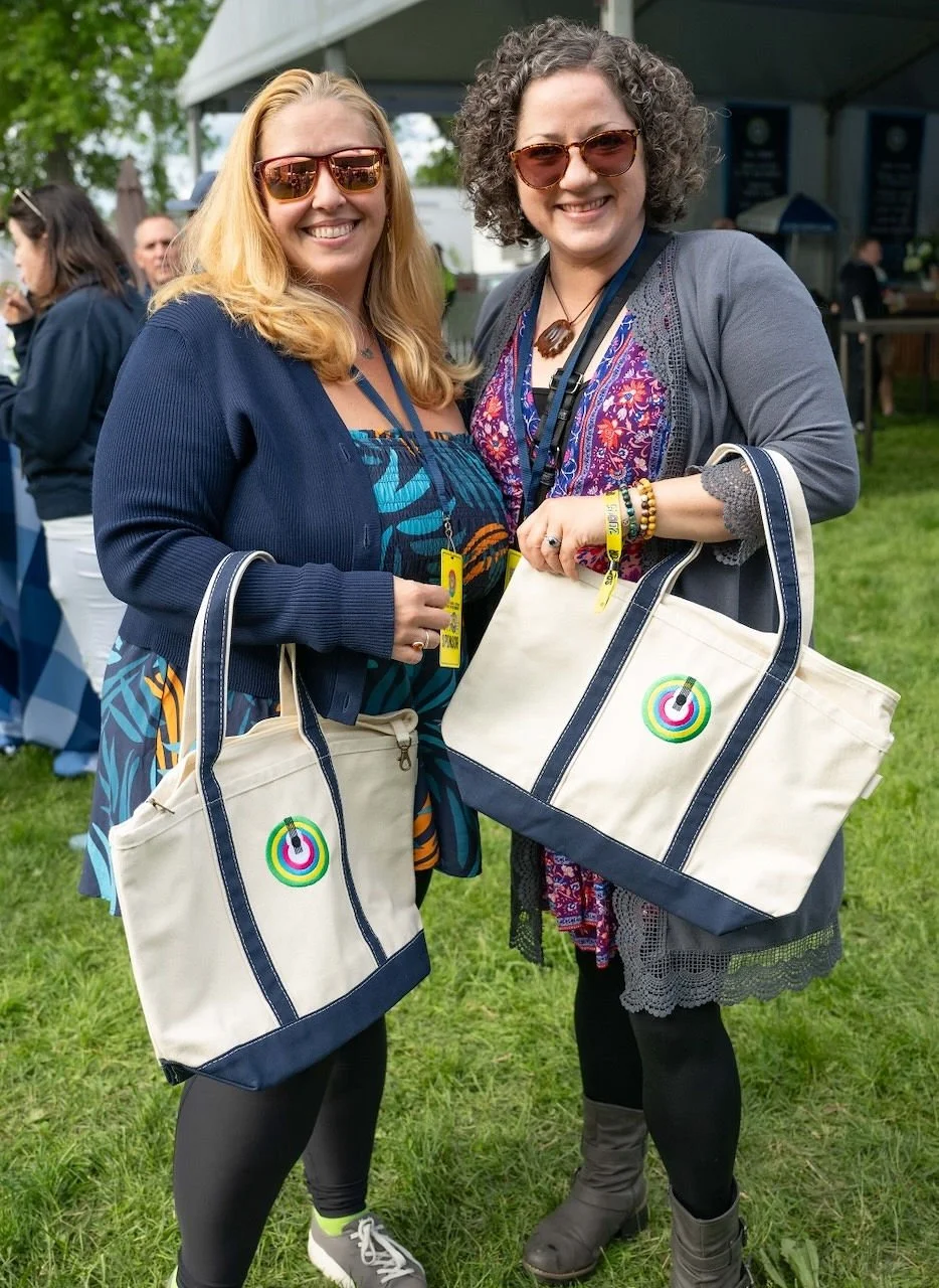 Two women smiling outdoors at an event, holding tote bags with colorful target circles on them. They are both wearing sunglasses and casual clothing, with other people and a tent in the background.