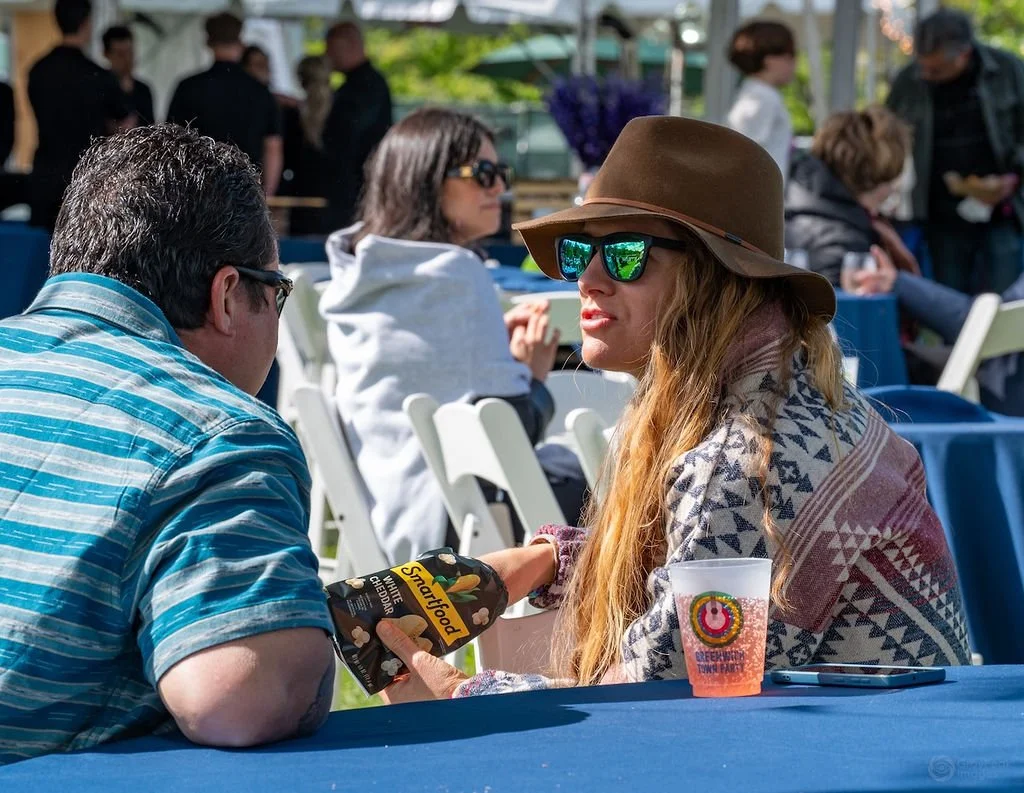 A woman wearing sunglasses, a wide-brimmed hat, and a patterned shawl sits at an outdoor event. She is engaging in a handshake with a man in a blue striped shirt. There are other people and tables in the background.