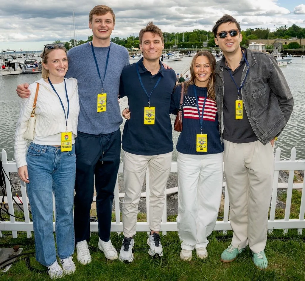 Group of five young adults standing outdoors near a body of water with boats and a cloudy sky in the background. They are smiling and wearing yellow sponsor badges around their necks.