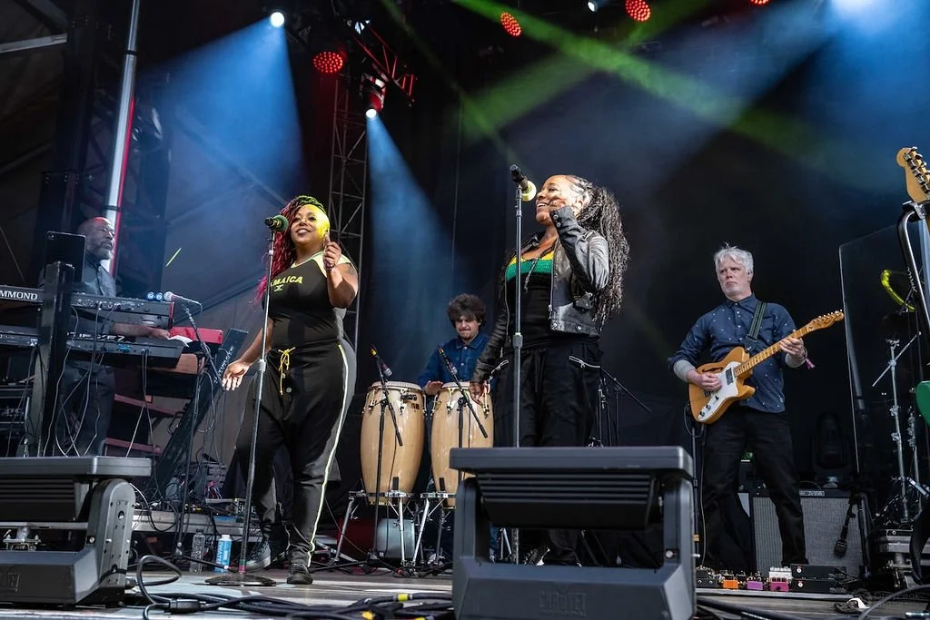 Band performing on stage with two female singers, guitarist, percussionist, and keyboardist, surrounded by stage equipment and colorful lights.