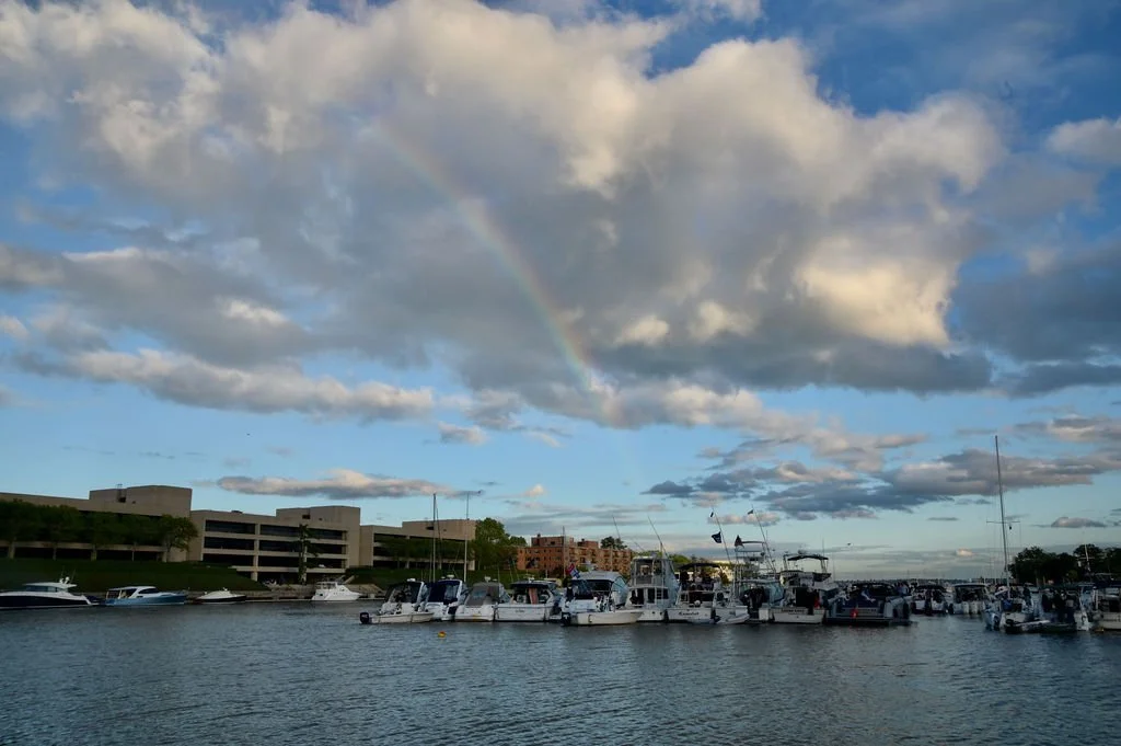 A harbor with boats docked, a cloudy sky with a rainbow, and a building in the background.