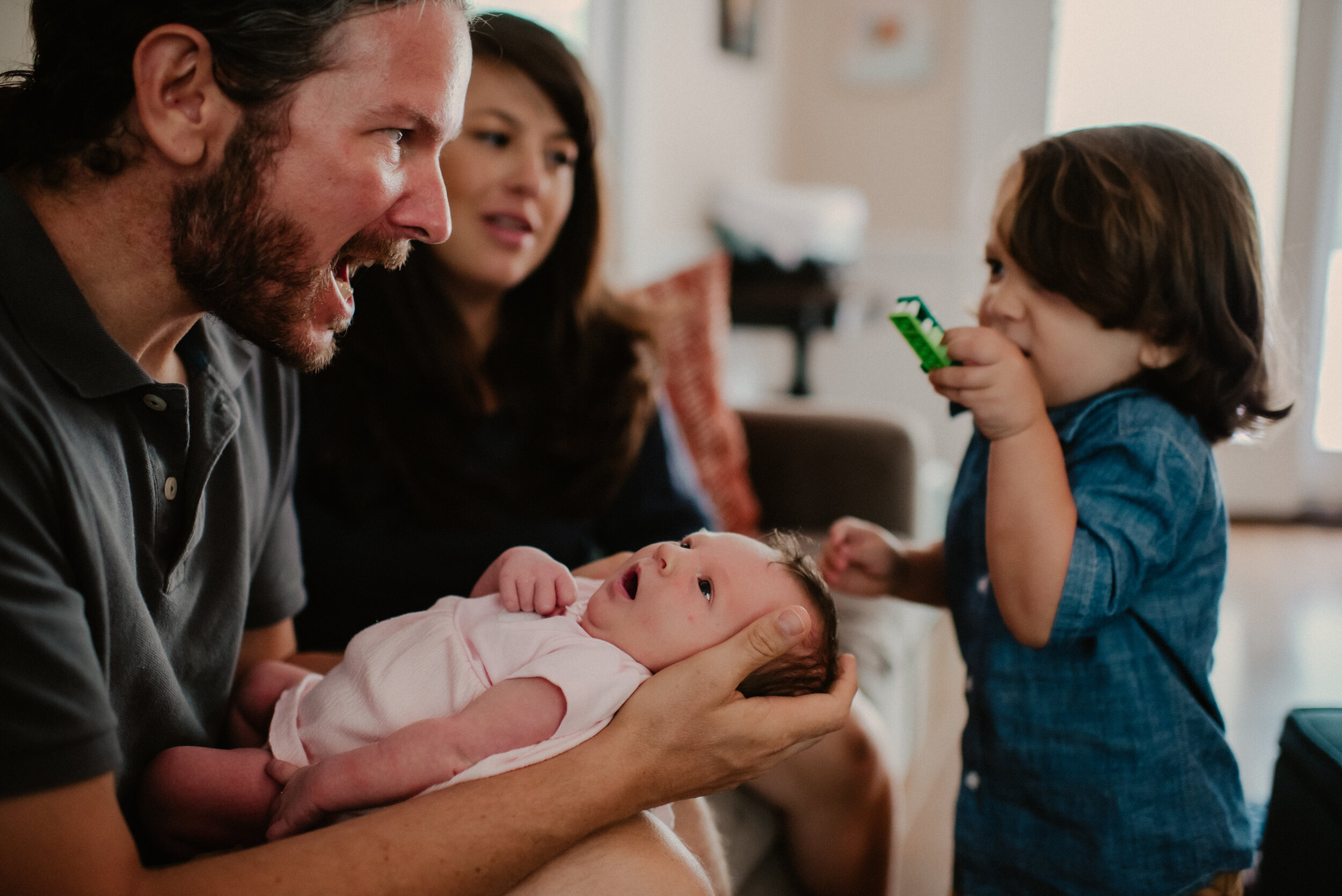 big brother shows green toy car to new baby sibling while dad holds newborn