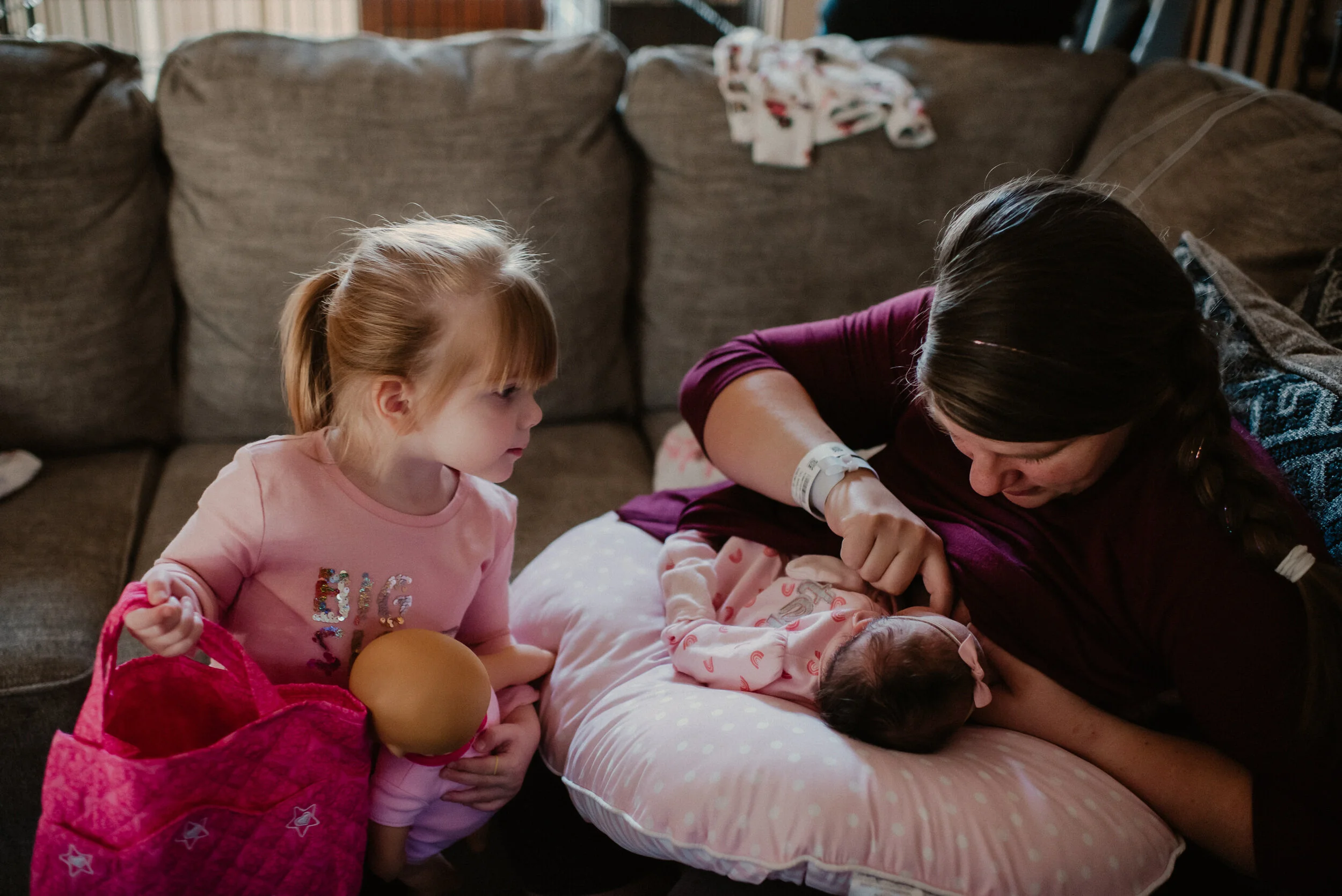 mother breastfeeds baby at home with curious big sister pretending to nurse her baby doll