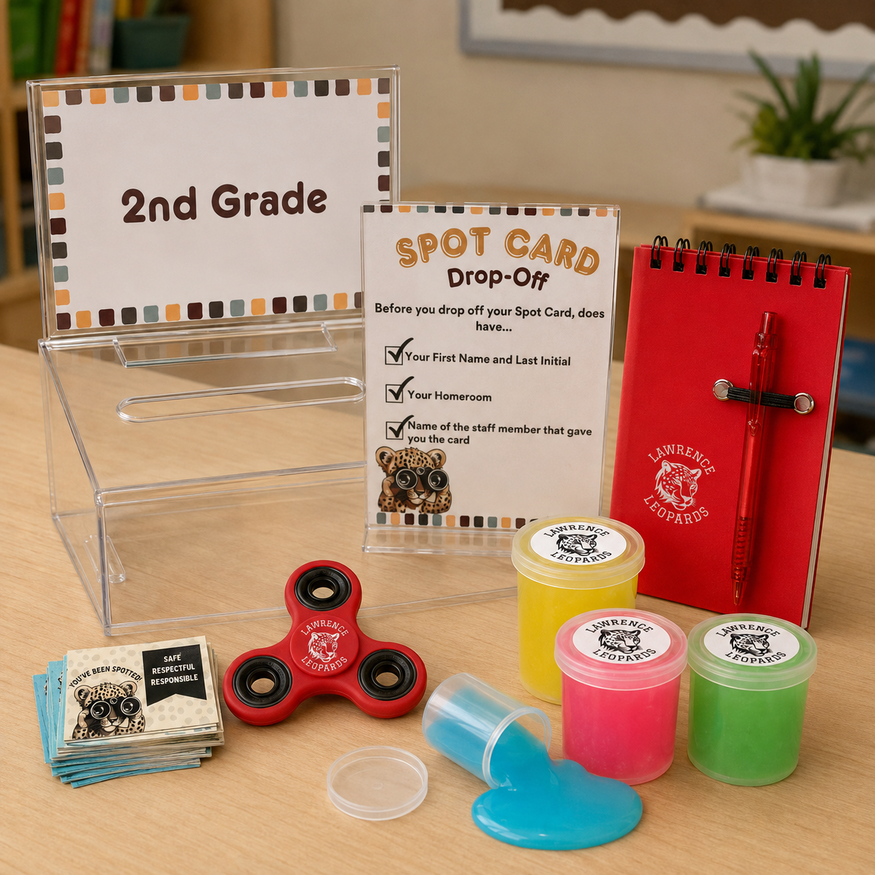 School classroom display with signs for 2nd grade and a drop-off spot card station, including slime supplies, a red fidget spinner, informational brochures about leopard safety, and a red Lawrence Leopards notebook with a pen.