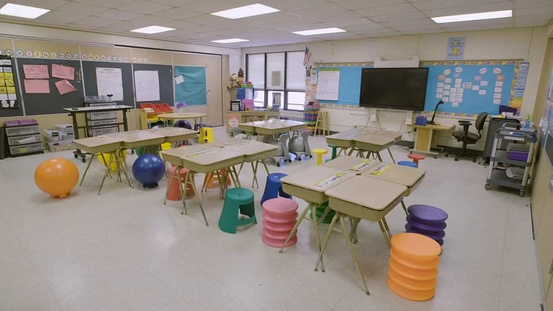 Classroom with desks, colorful stools, exercise balls, educational posters, a large screen, and supplies.