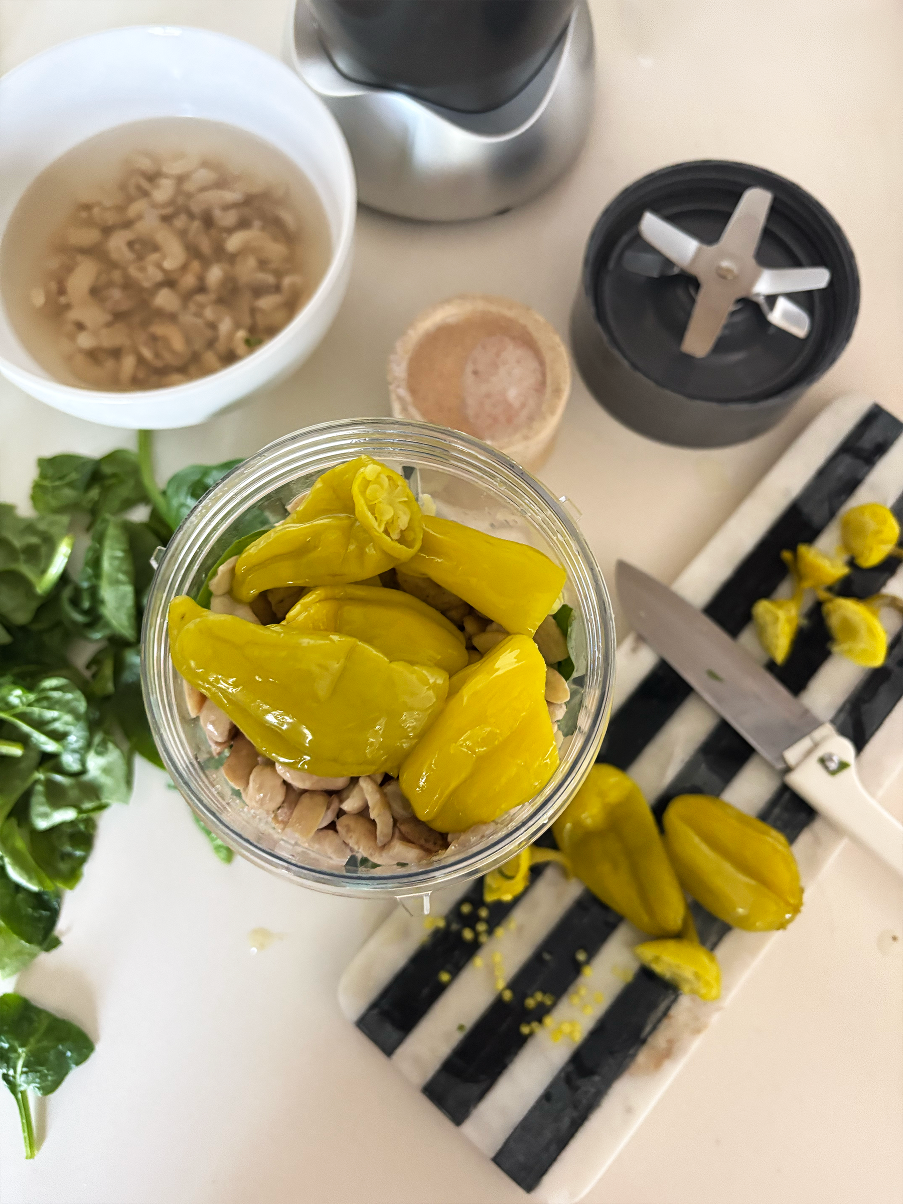 Top view of white kitchen counter with pepperocinis on black and white striped cutting board, fresh spinach and basil, sea salt, and mixed ingredients in nutribullet blender