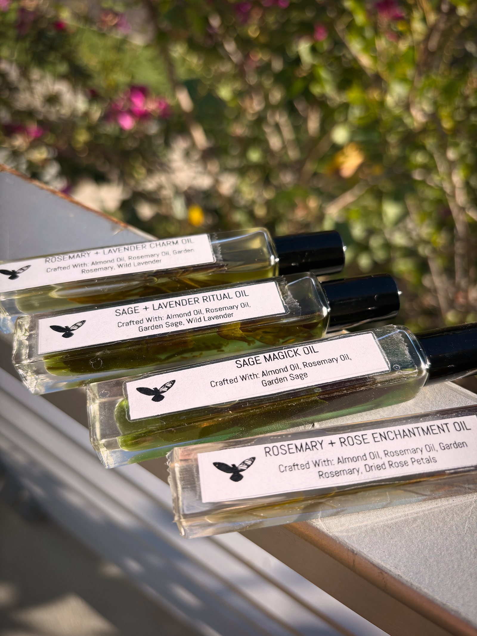 Four rosemary oils on ledge with greenery in background, all branded with the bird and the butterfly logo and description of ingredients