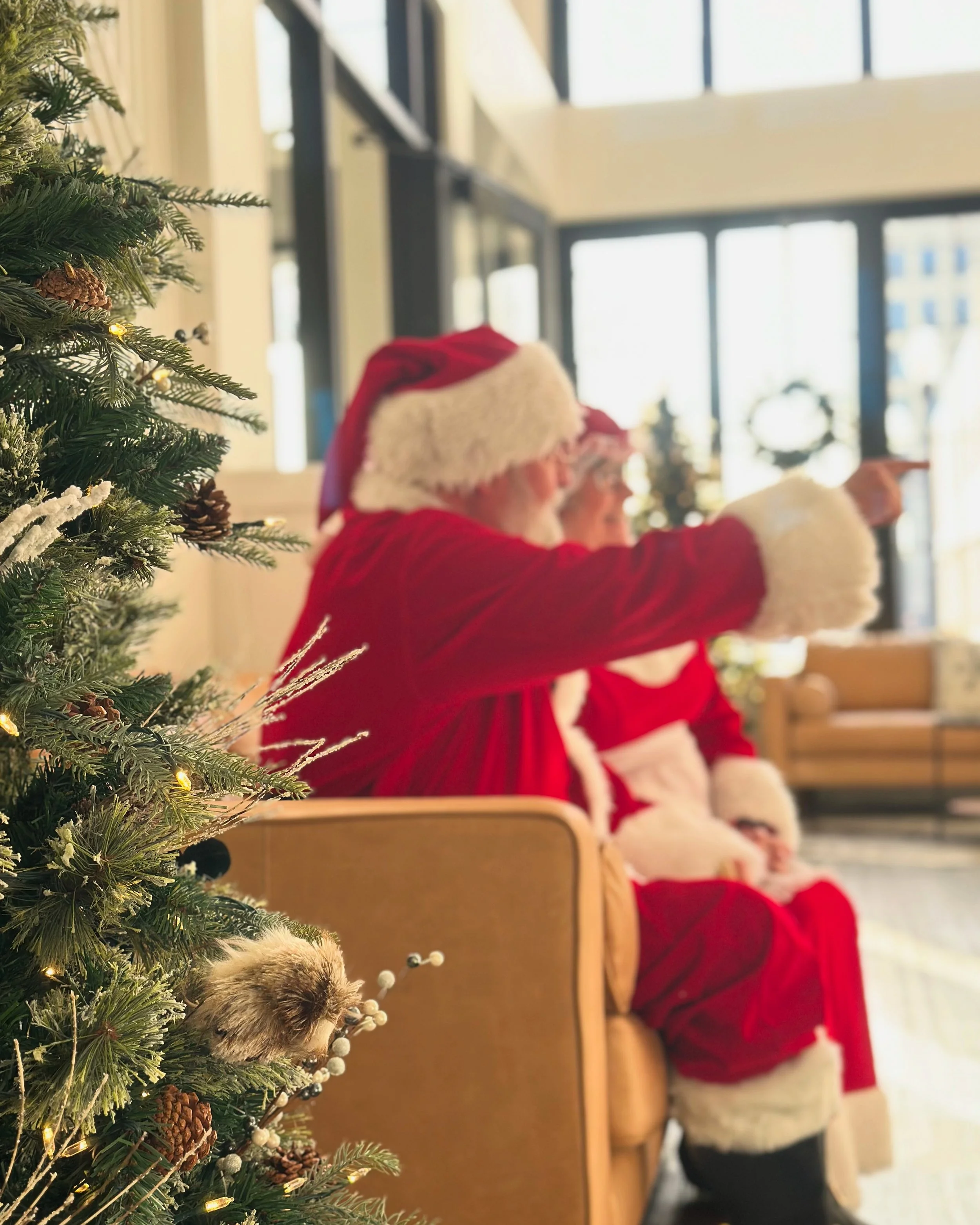 Children in Santa costumes sitting indoors near a decorated Christmas tree, with one child pointing toward something.