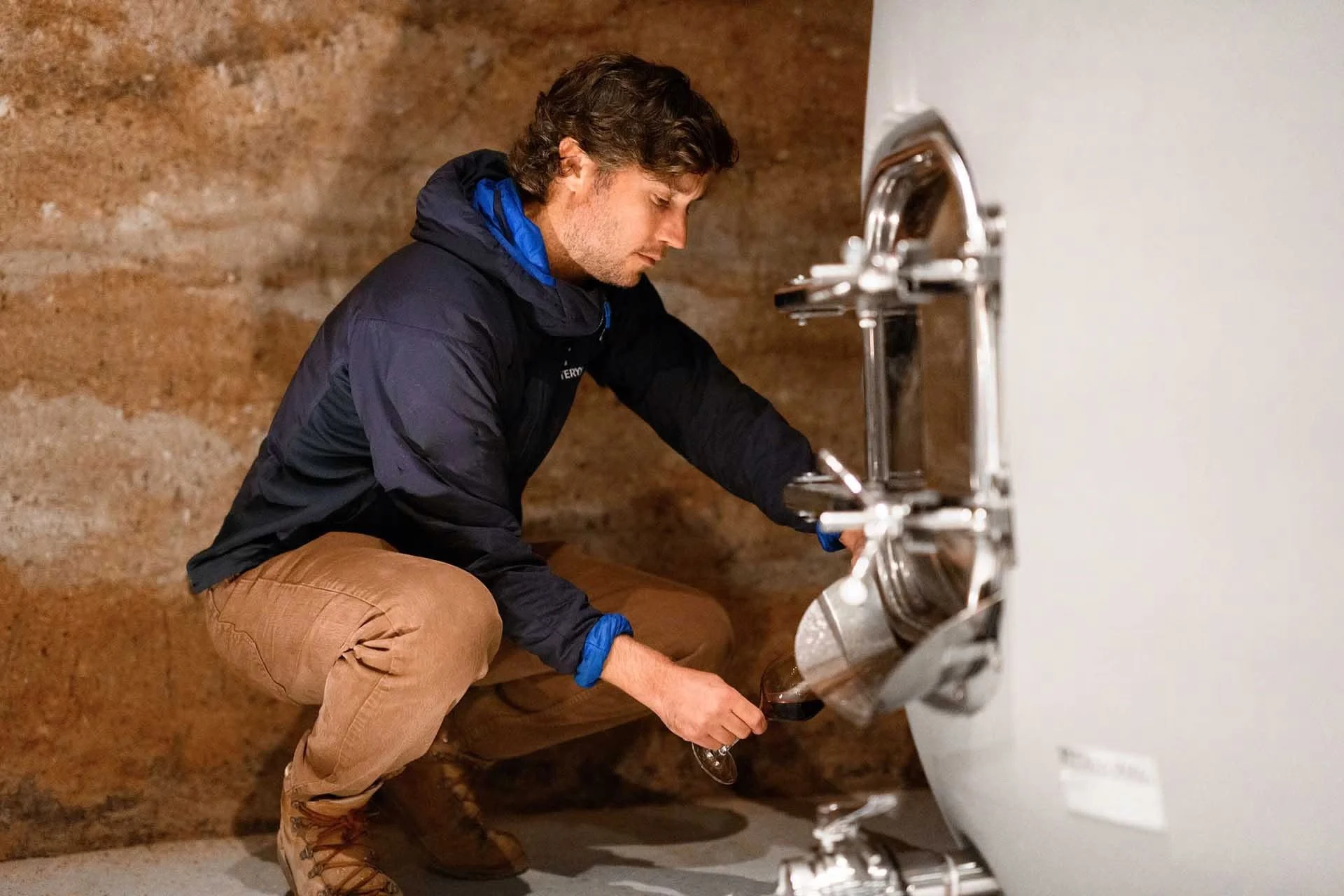 A man in outdoor clothing is crouching and pouring wine from a tap into a glass.