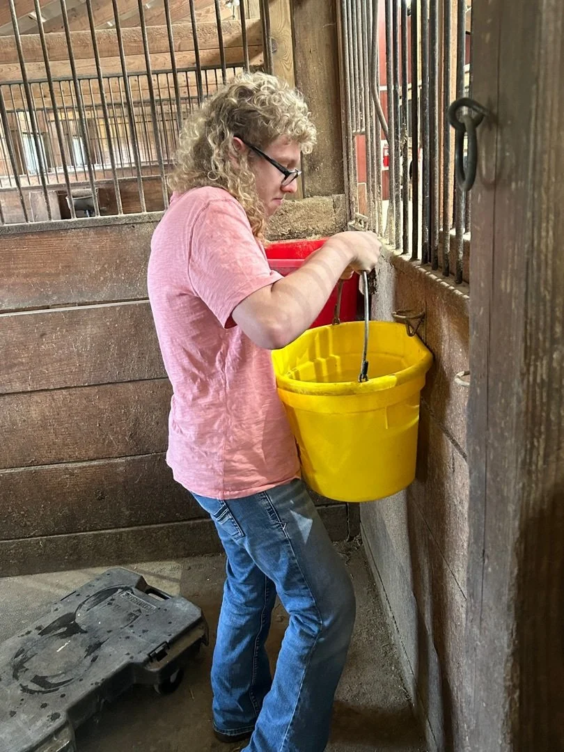A People with Purpose participant holding up a yellow bucket of water in a horse barn