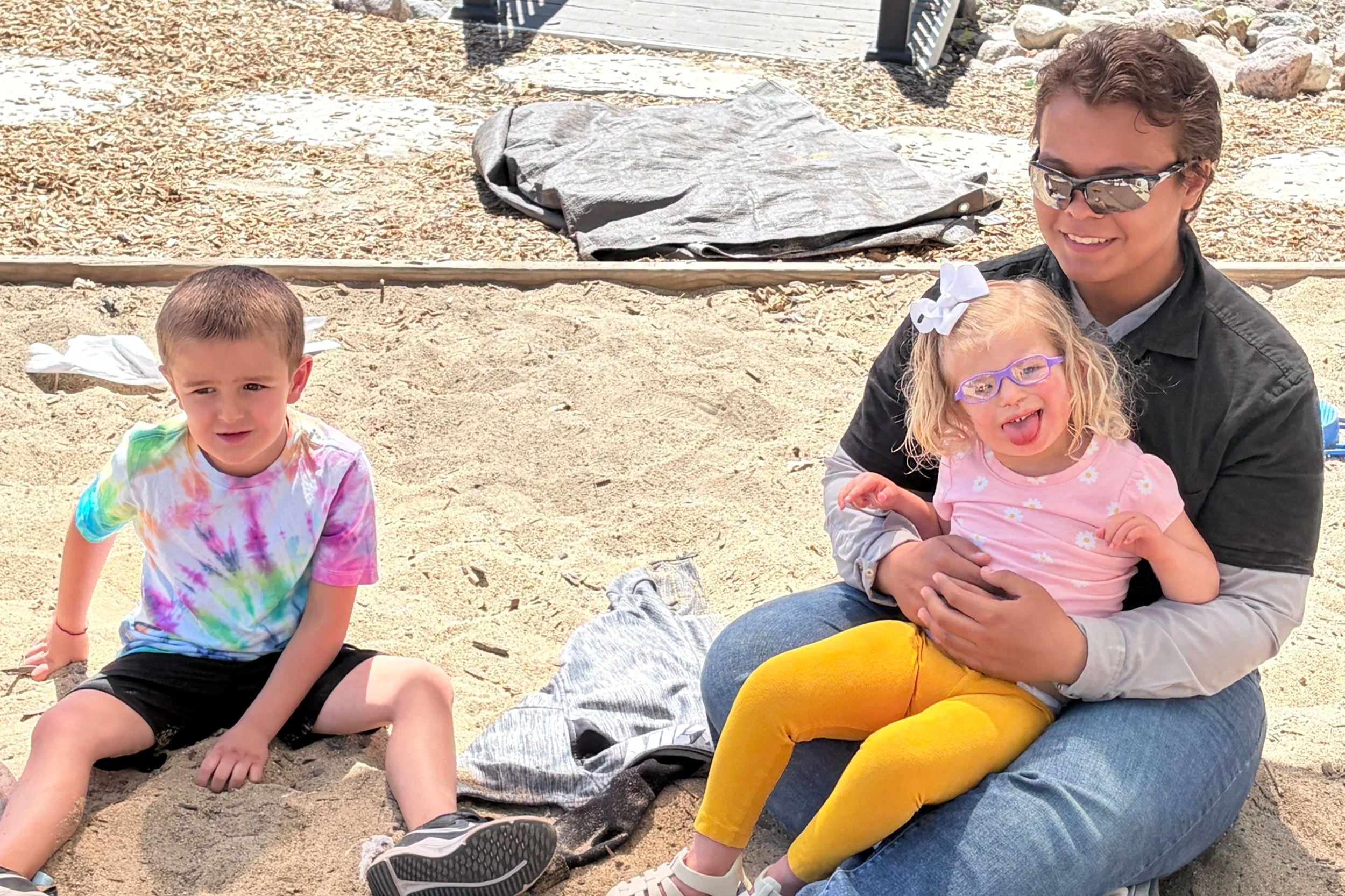 Two children and a teacher sitting in a sandbox