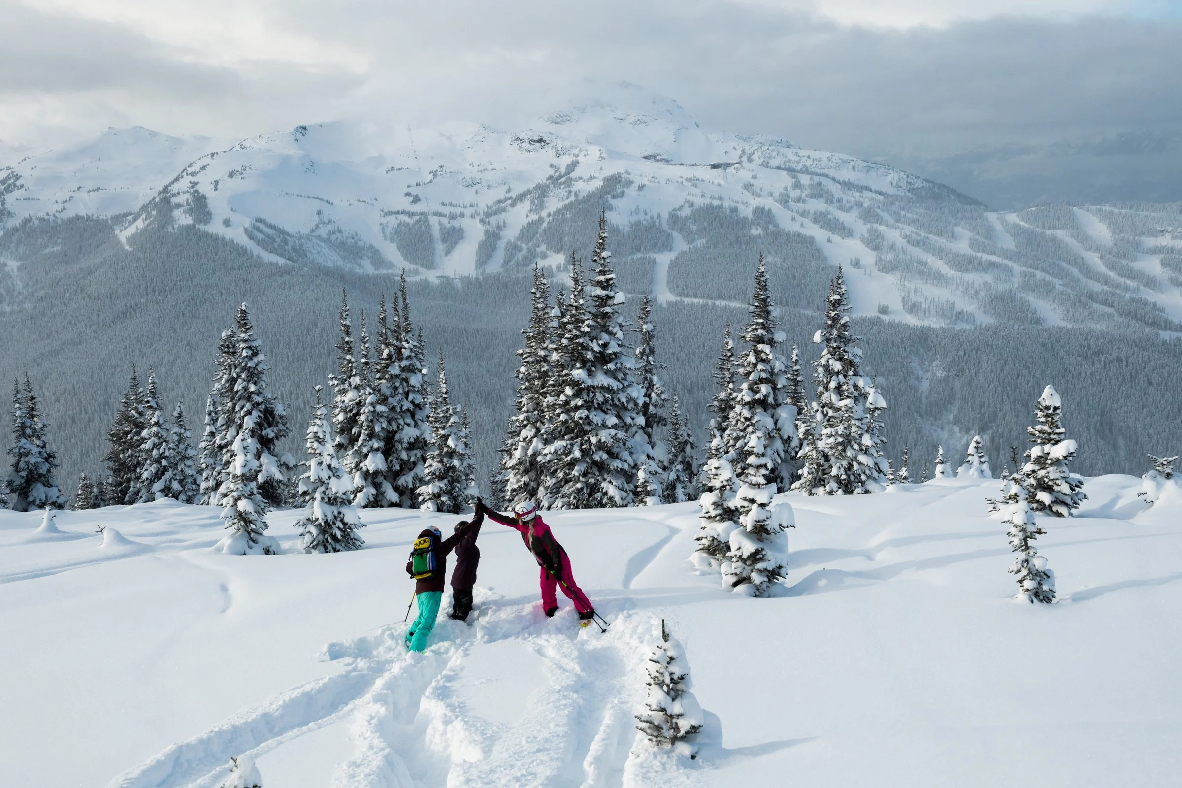 Young friends playing in a snowy day at Whistler Blackcomb resort with pine trees and ski trails in the background under cloudy skies.